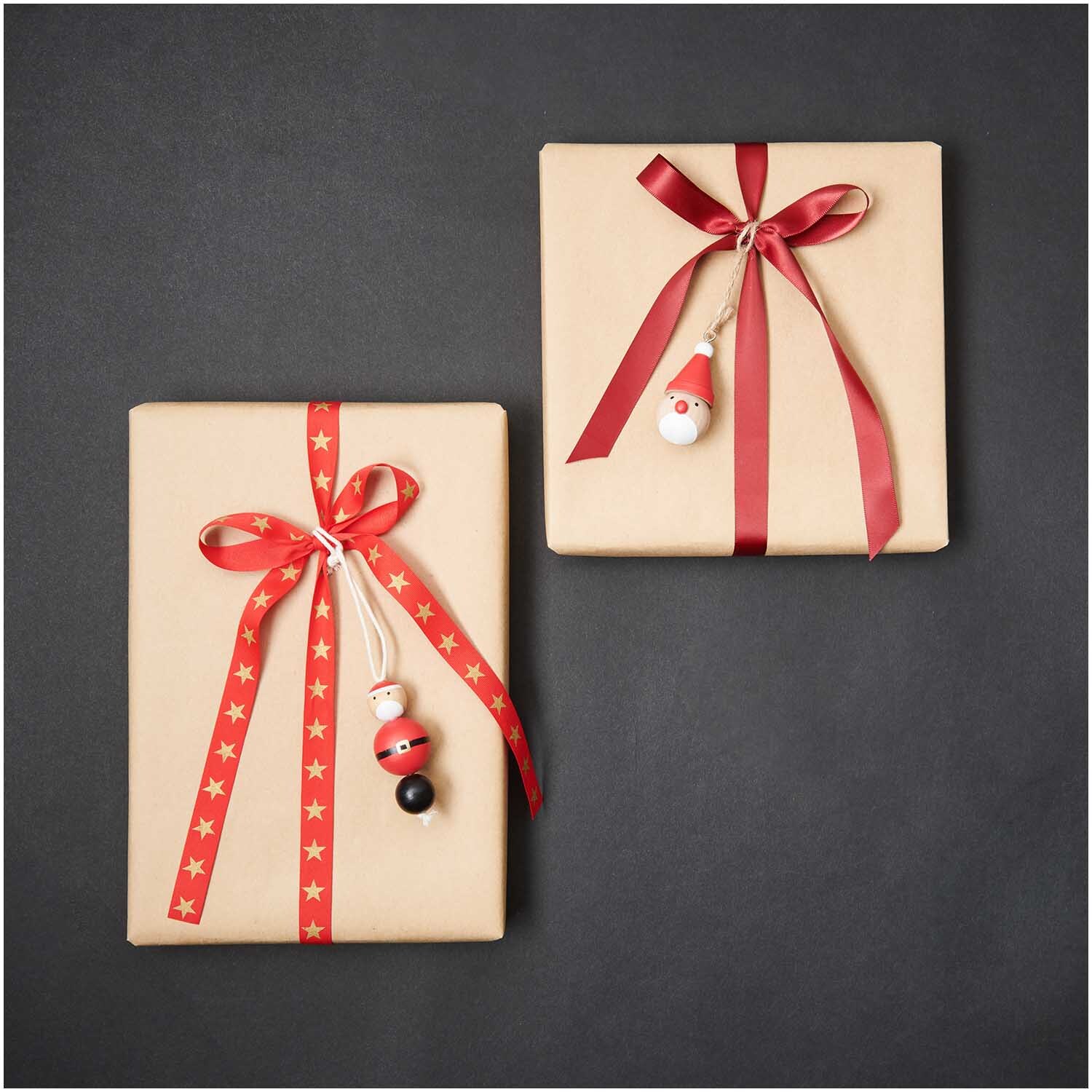 Holzhänger Santa 3x6,5cm von Rico Design an zwei Geschenken mit roten Schleifen auf schwarzem Hintergrund.