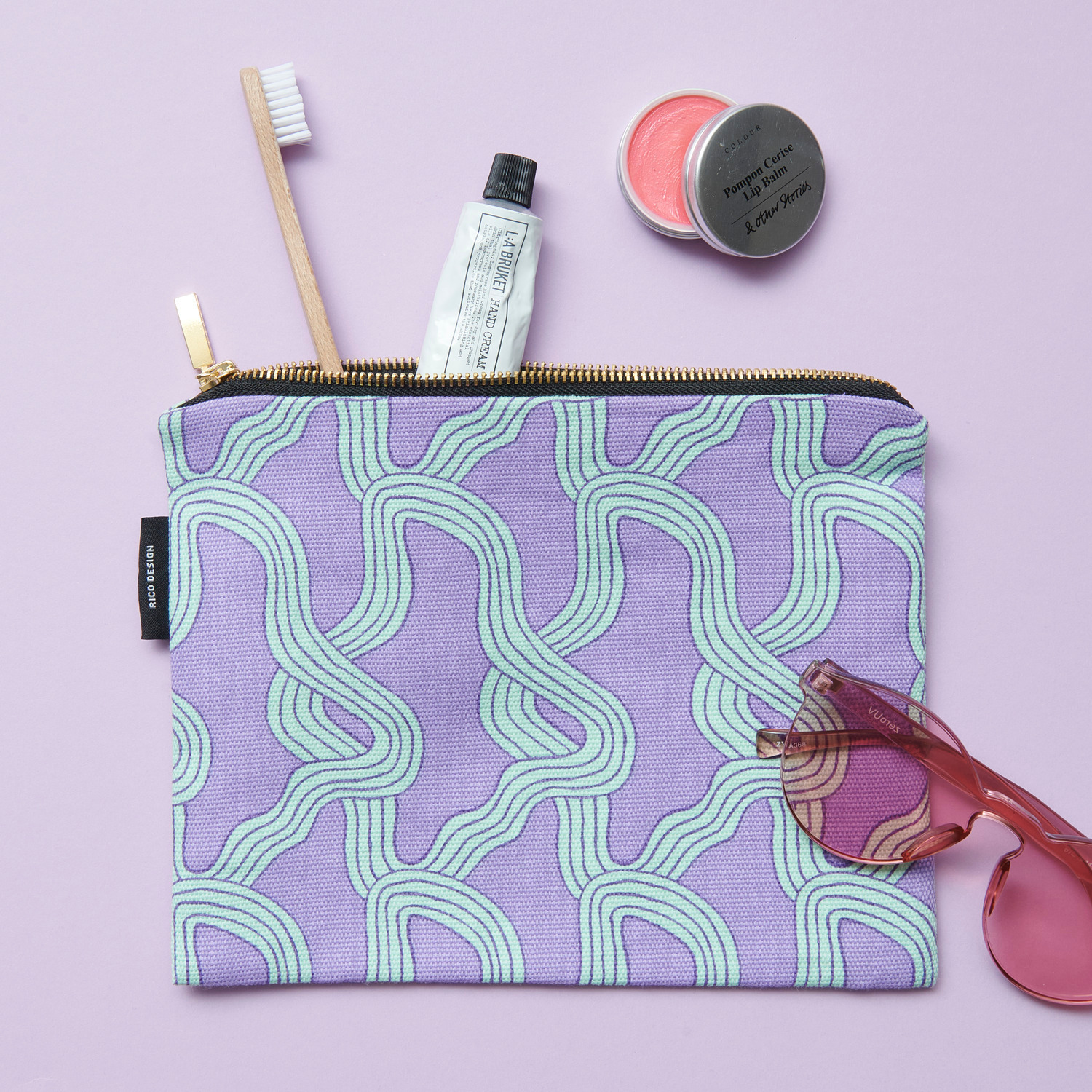 Canvas-Pouches Myzel Flieder 17x23cm von Rico Design Canvas-Pouches Myzel Flieder: Lila Tasche mit geschwungenem Muster, Zahnbürste, Creme und Sonnenbrille.