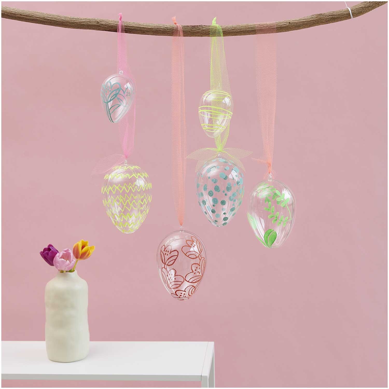 Glas Eier transparent 4 Stück Ø 3cm von Rico Design Glas Eier transparent 4 Stück Ø 3cm, dekorativ hängend an einem Ast vor rosa Hintergrund.