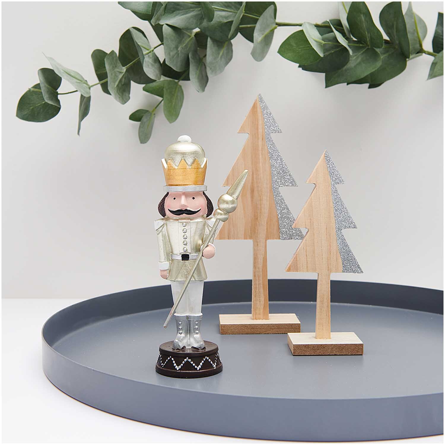 Polyresin Figur Nussknacker mit Stab 17cm neben zwei dekorativen Holzbäumen auf einem Tablett.