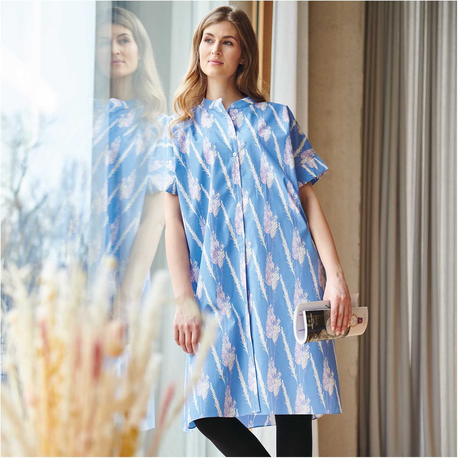 Meterware Druckstoff Pampasgras azur von Rico Design Meterware Druckstoff Pampasgras azur: Frau in azurblauem Kleid mit Pampasgras-Muster, steht am Fenster.