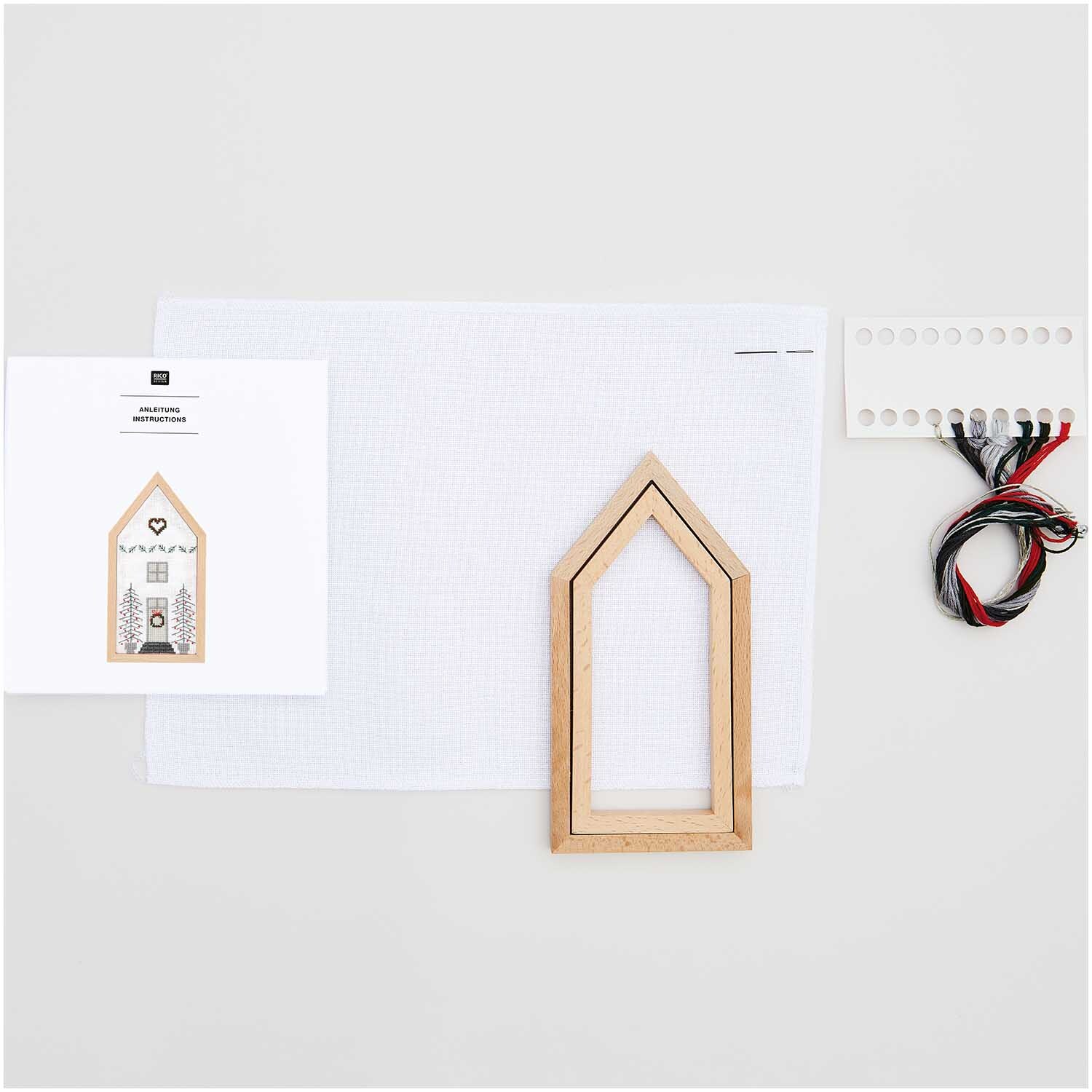 Paquet de broderie Maison petite 9x18cm from Rico Design Stickpackung Haus klein 9x18cm von Rico Design mit Holzrahmen, Stoff, Anleitung und Garn auf hellem Hintergrund.