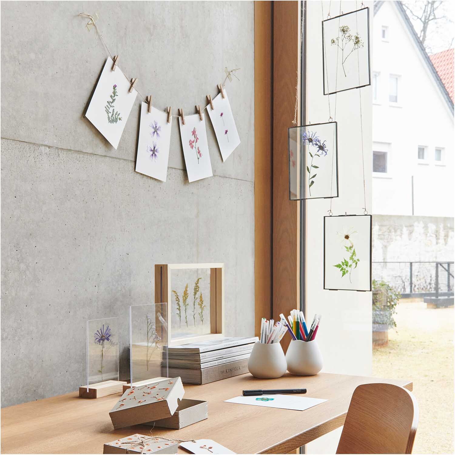 Holzaufsteller mit doppelter Acrylscheibe 10x15cm Farbe Weiß von Rico Design Holzaufsteller mit doppelter Acrylscheibe 10x15cm von Rico Design auf einem Schreibtisch mit Pflanzenmotiven und Schreibutensilien.