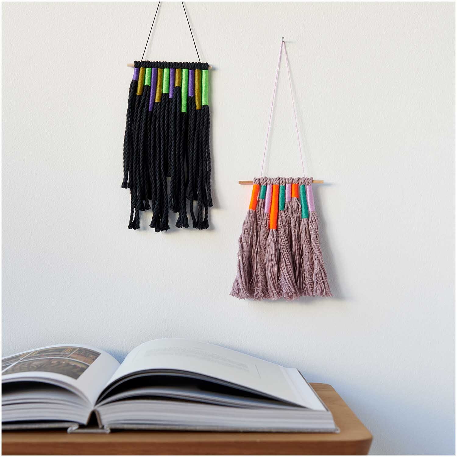 Set de fils à broder Multicolour Fashion 24 pièces 24-teilig from Rico Design Stickgarn Set Multicolour Fashion: Zwei Wandbehänge aus buntem Garn, dekorativ über einem offenen Buch.