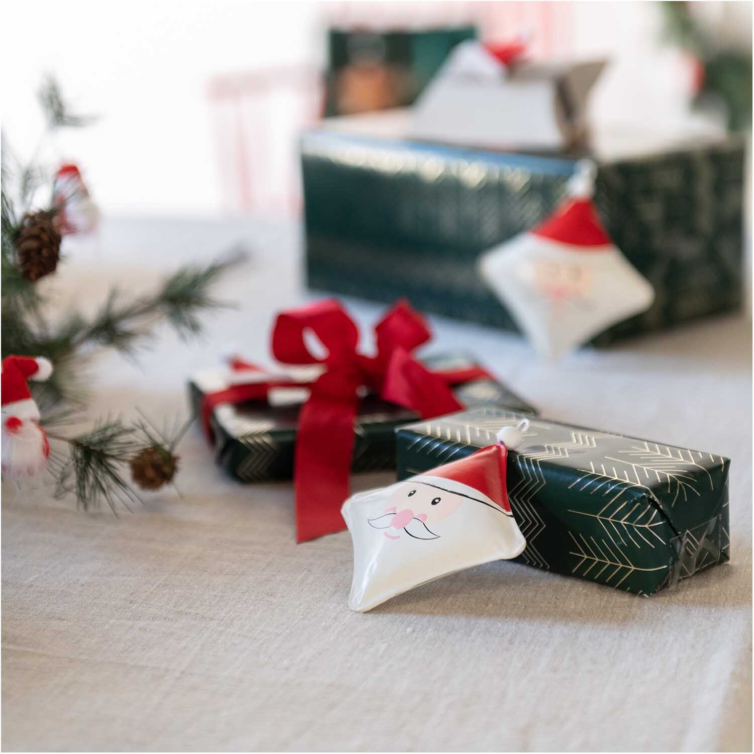 Metallhänger Weihnachtsmann rot-weiß 10x10cm von Rico Design Metallhänger Weihnachtsmann rot-weiß 10x10cm, dekorativ auf Geschenken arrangiert, umgeben von Tannenzweigen.