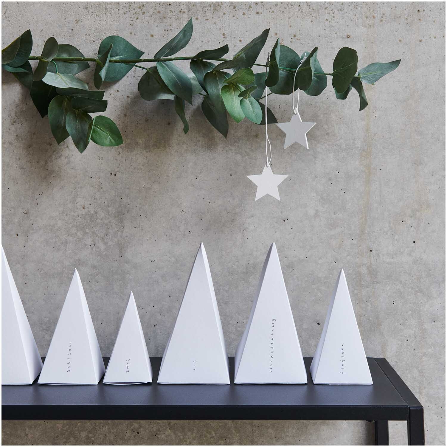 Paper Poetry Adventskalender Bäume 24 Stück Farbe Weiß von Rico Design Paper Poetry Adventskalender Bäume 24 Stück, weiße Pyramiden auf schwarzem Regal vor Betonwand.