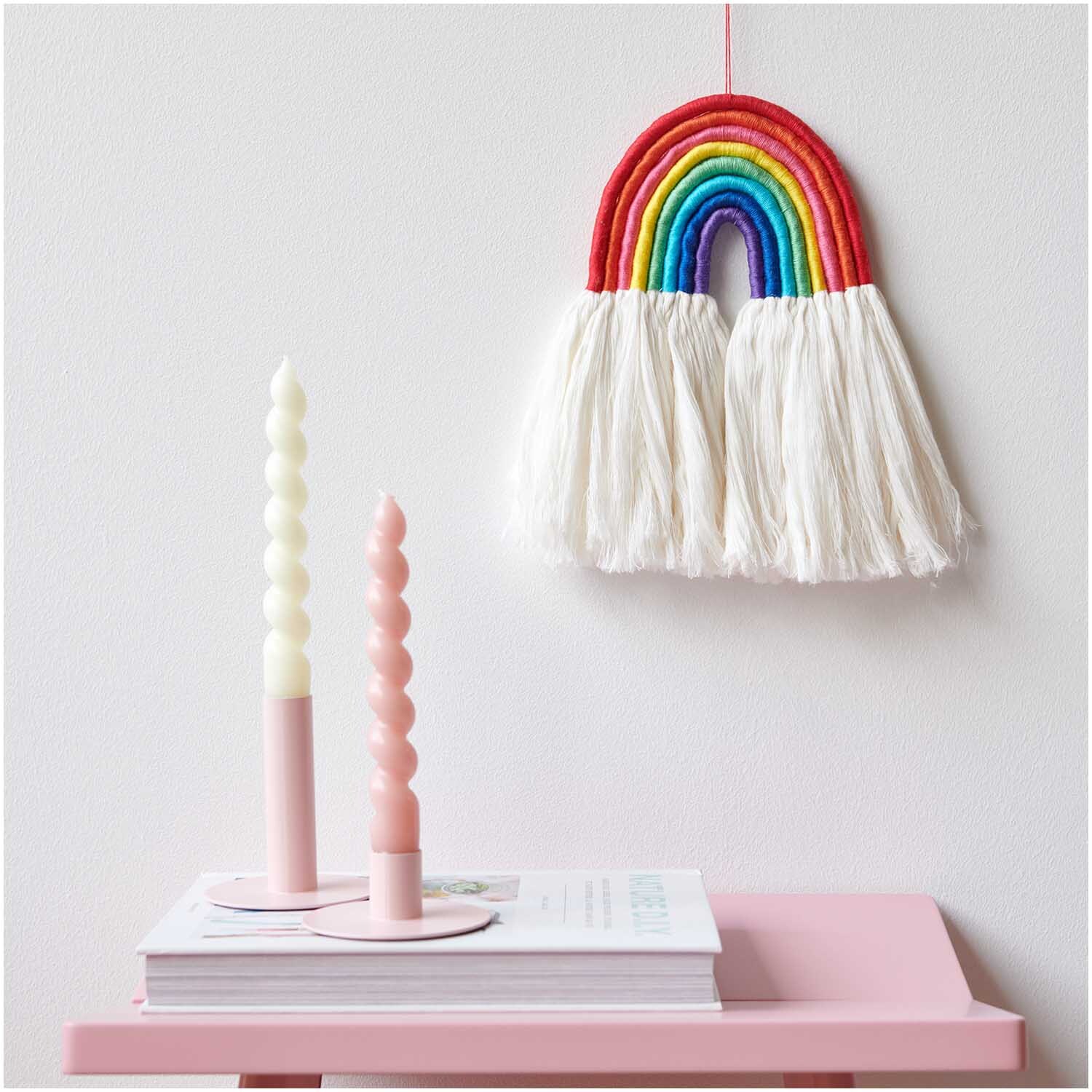 Set de fils à broder Rainbow 10 pièces 10-teilig from Rico Design Stickgarn Set Rainbow, 10-teilig: Bunte Wanddekoration in Regenbogenform über rosa Tisch mit Kerzen.