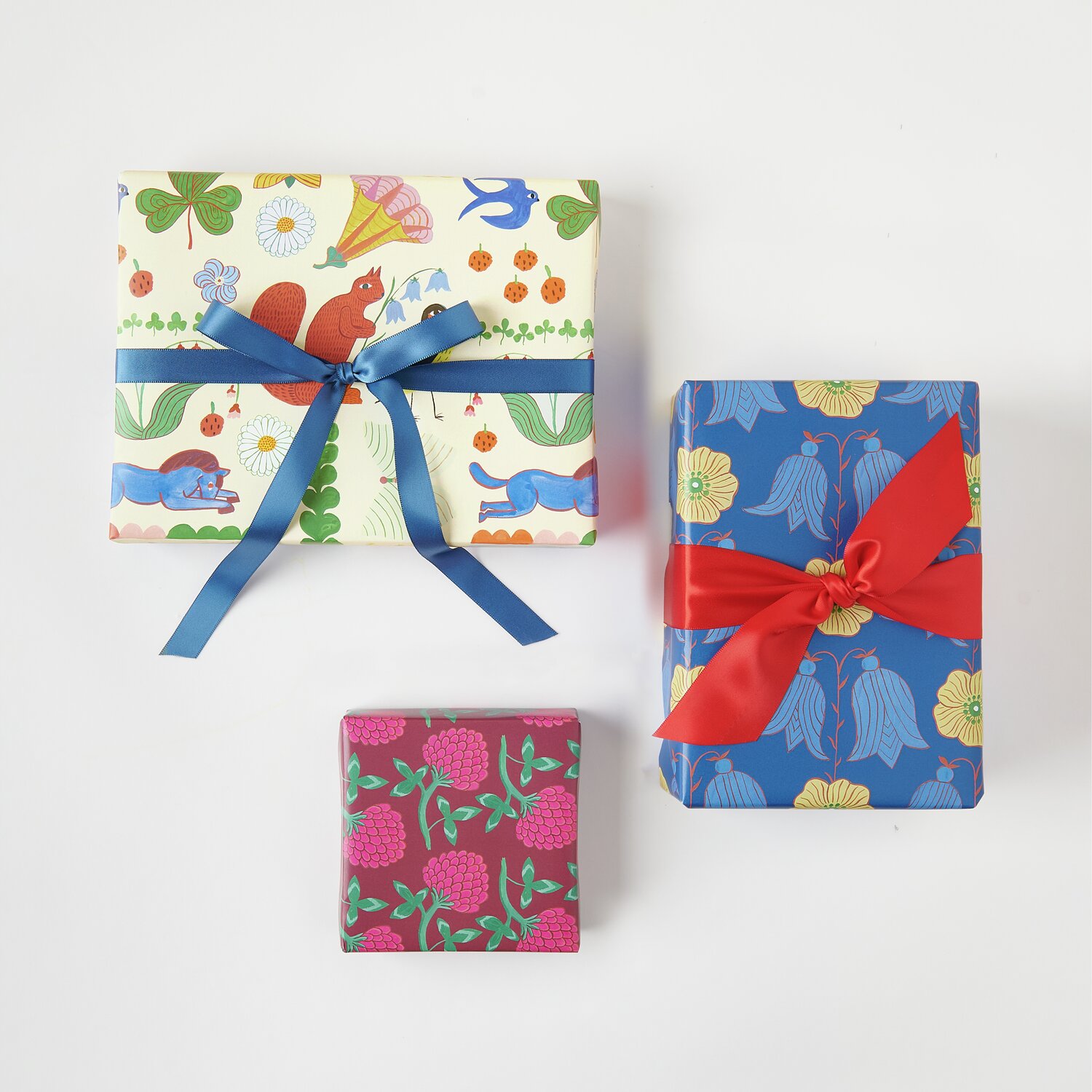 Geschenkpapier Rotklee 70cm 2m 100g/m² von Rico Design Geschenkpapier Mix Midsommar Magic: Drei Geschenke in buntem Papier mit blauen und roten Schleifen.