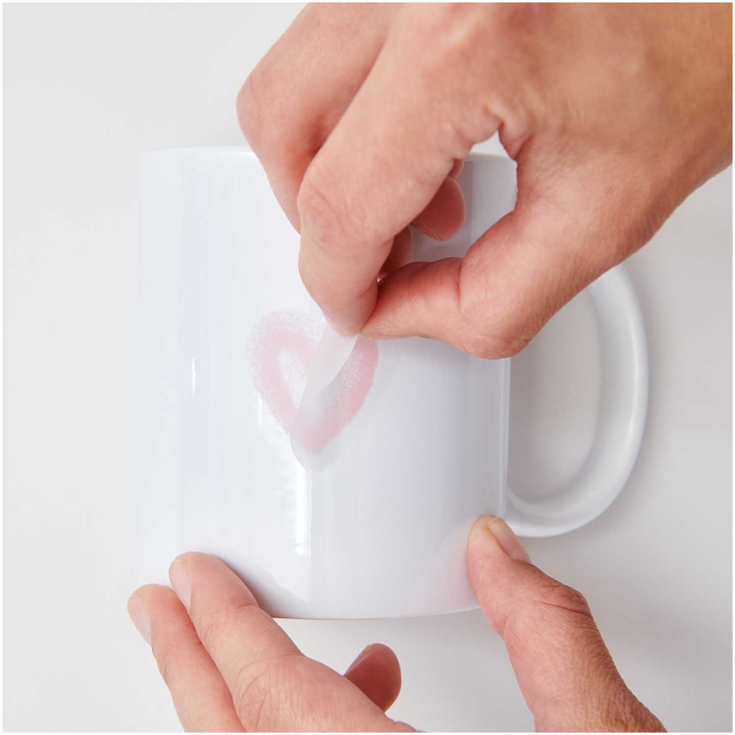 Transfermotiv Spray Herzen 12 Stück von Rico Design Transfermotiv Spray Herzen: Hände kleben ein Herzmotiv auf eine weiße Tasse. Marke: Rico Design.