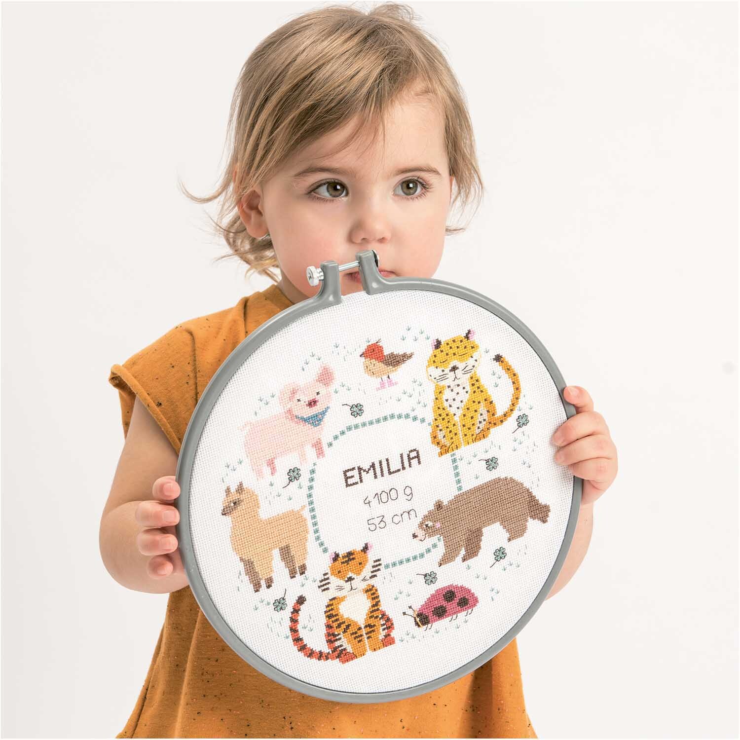 Paquet à broder Image de naissance animaux from Rico Design Stickpackung Geburtsbild Tiere von Rico Design zeigt ein Kind mit einem bestickten Rahmen voller Tiermotive.