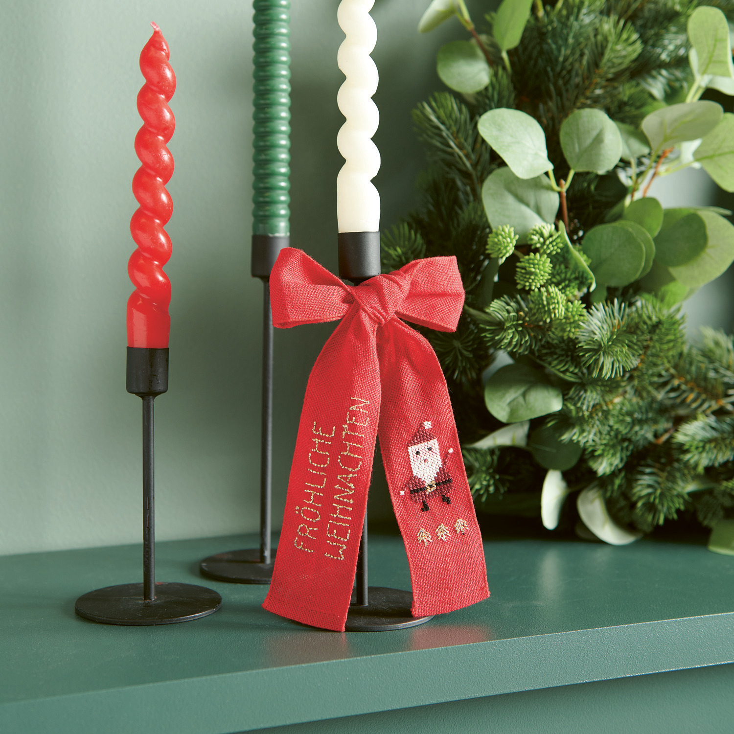 Stickpackung Schleife Nikolaus 12x19cm von Rico Design Stickbuch Put a Bow on It Nr. 185: Rote Schleife mit Nikolausmotiv, dekoriert neben Kerzen auf grünem Regal.