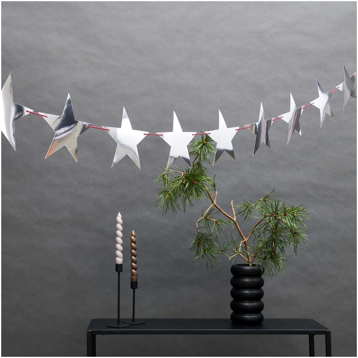 Paper Poetry Girlande Sterne silber 3m von Rico Design Paper Poetry Girlande Sterne silber 3m, dekorativ über einem Tisch mit Pflanzen und Kerzen.