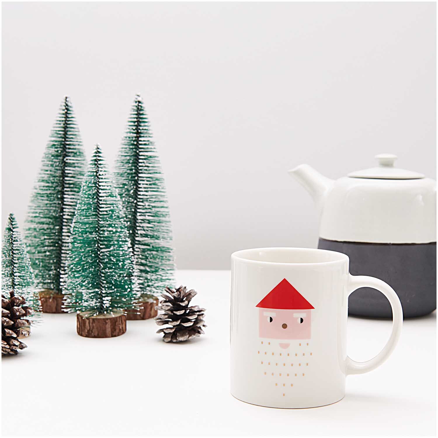 Porzellan Tasse Santa 8x9,5cm von Rico Design Porzellan Tasse Santa 8x9,5cm mit rotem Santa-Motiv, neben grünen Miniaturtannen und weißer Teekanne.