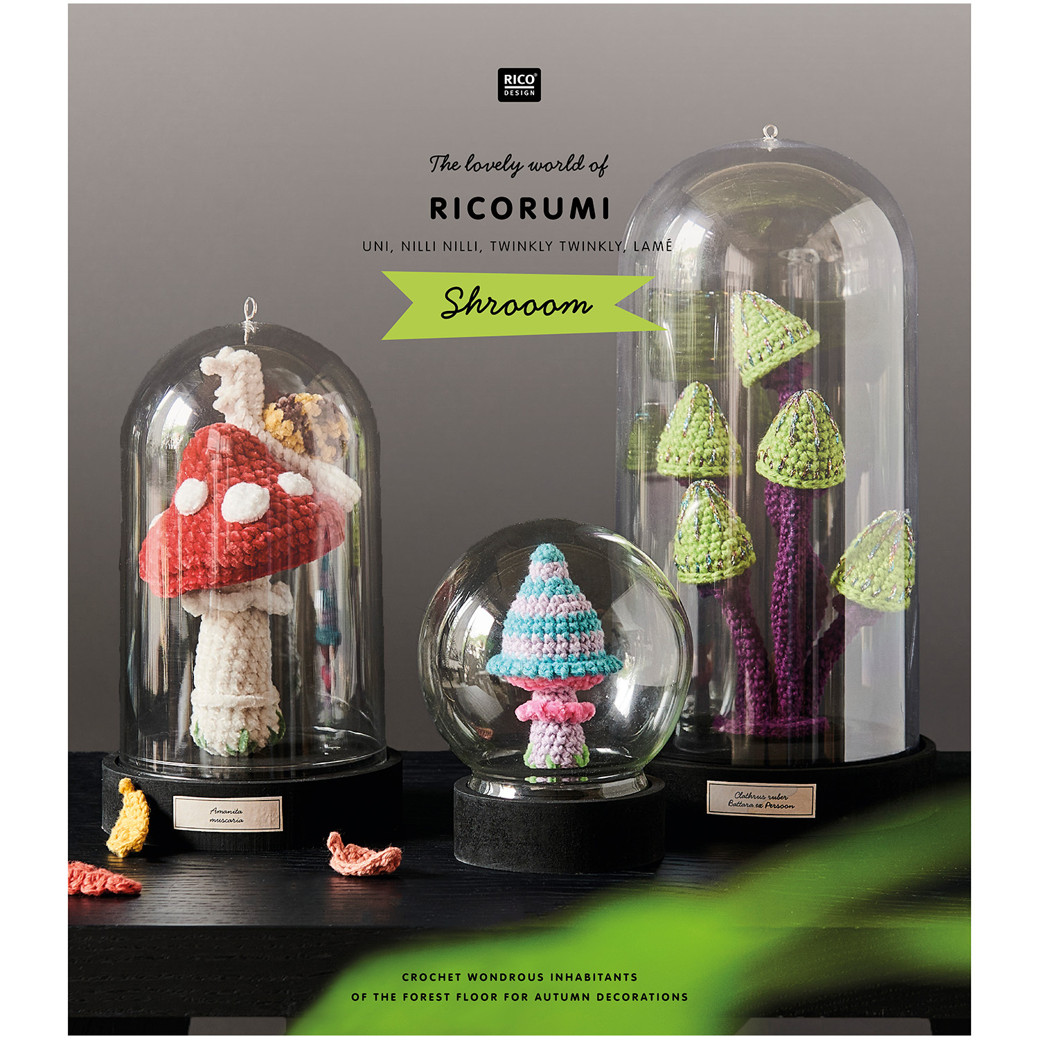 Ricorumi Shrooom Sprachauswahl englisch von Rico Design Ricorumi Shrooom: Drei gehäkelte Pilze in Glaskuppeln, präsentiert von Rico Design, auf einem Tisch.