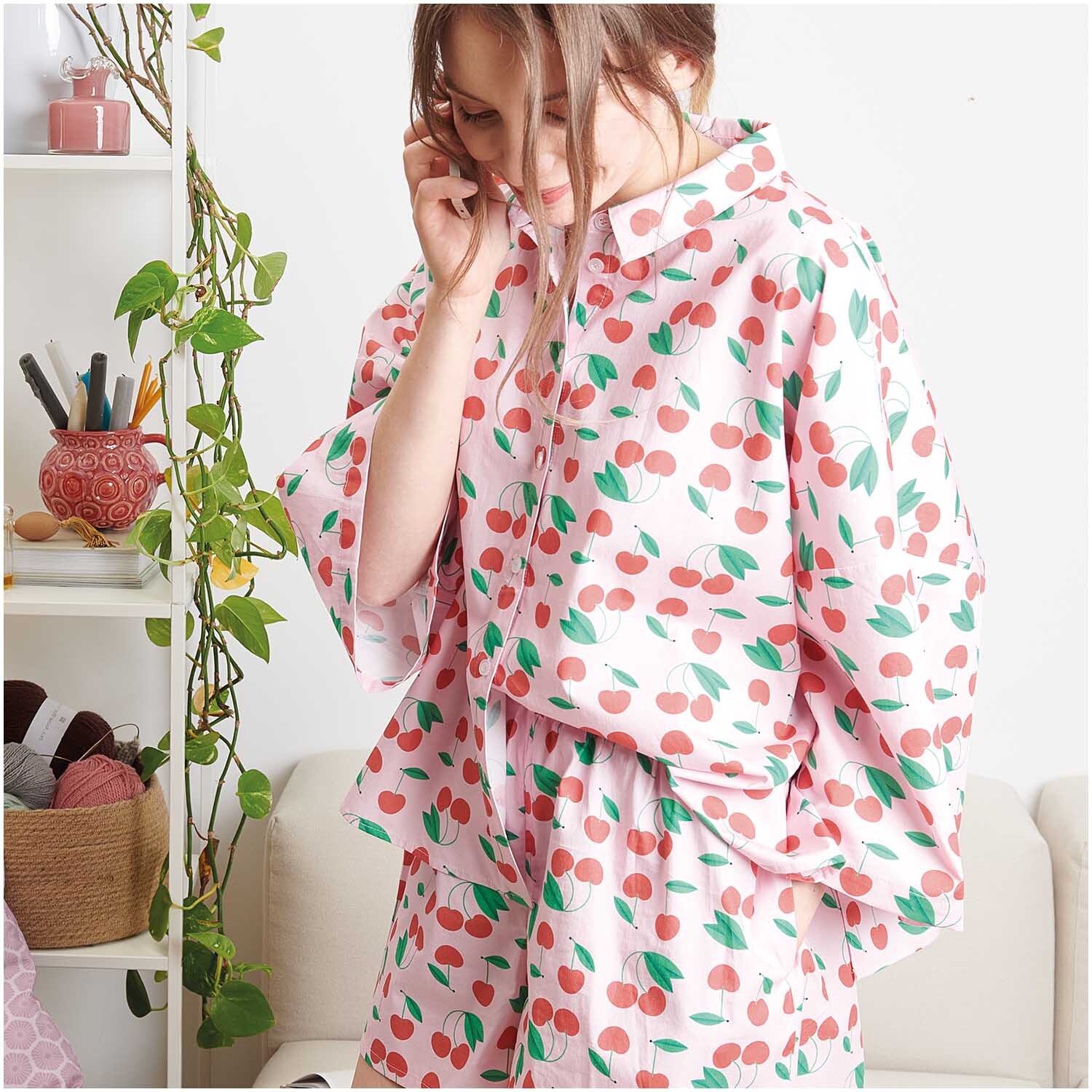 Stoffabschnitt Baumwoll-Popelin rosa Kirschen 50x140cm: Frau trägt Pyjama mit Kirschmuster, umgeben von Pflanzen und Garn.