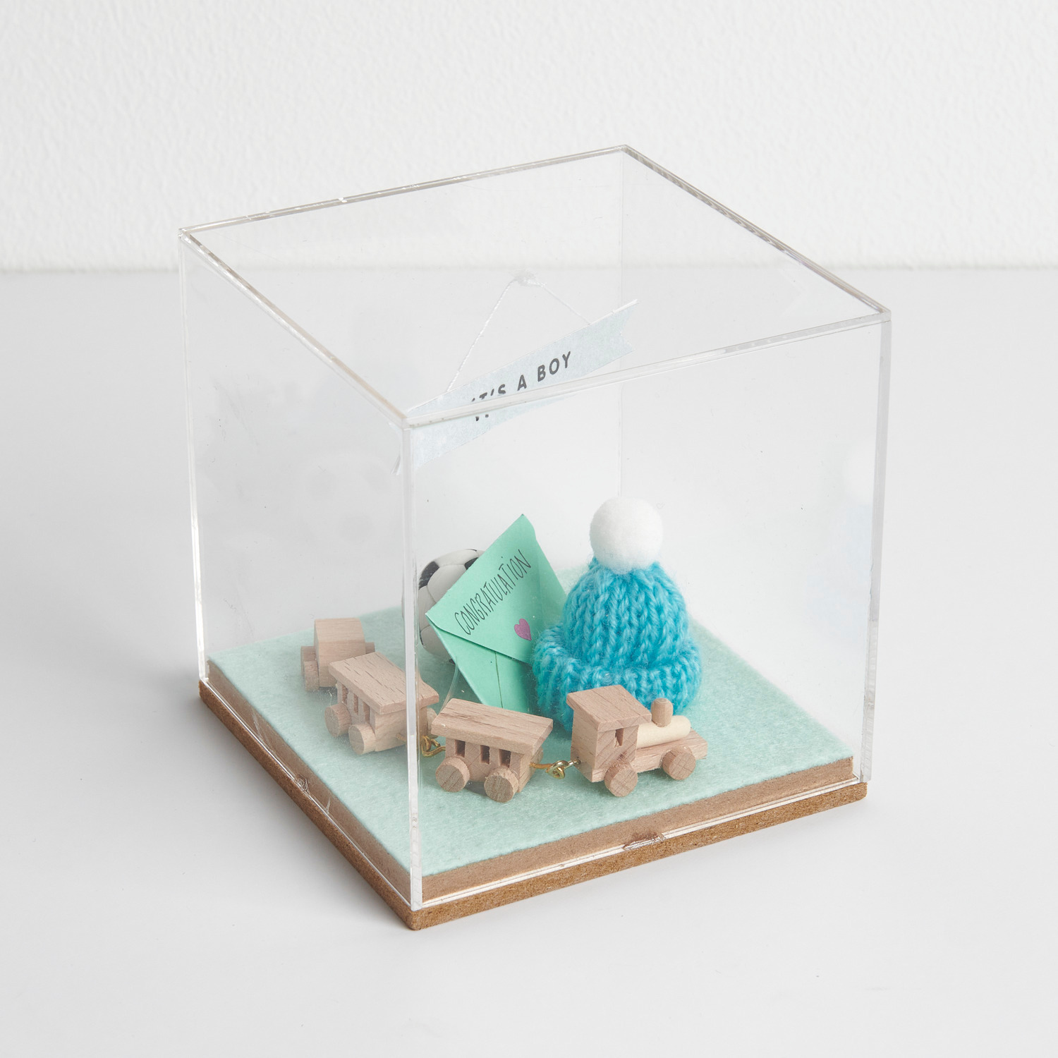Figurico Dekohaube Würfel Größe 6x6x6,5cm von Rico Design Figurico Dekohaube Würfel mit Holzspielzeug und blauem Strickmütze auf grünem Filzboden.