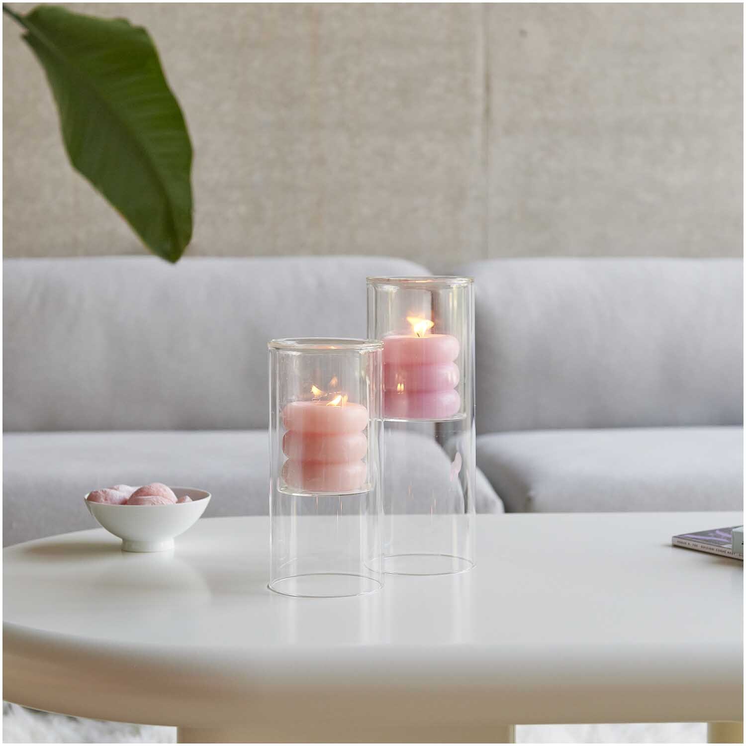 Glas-Windlicht mit brennender Kerze auf weißem Tisch, dekoriert mit rosa Elementen, Ø9,5cm.