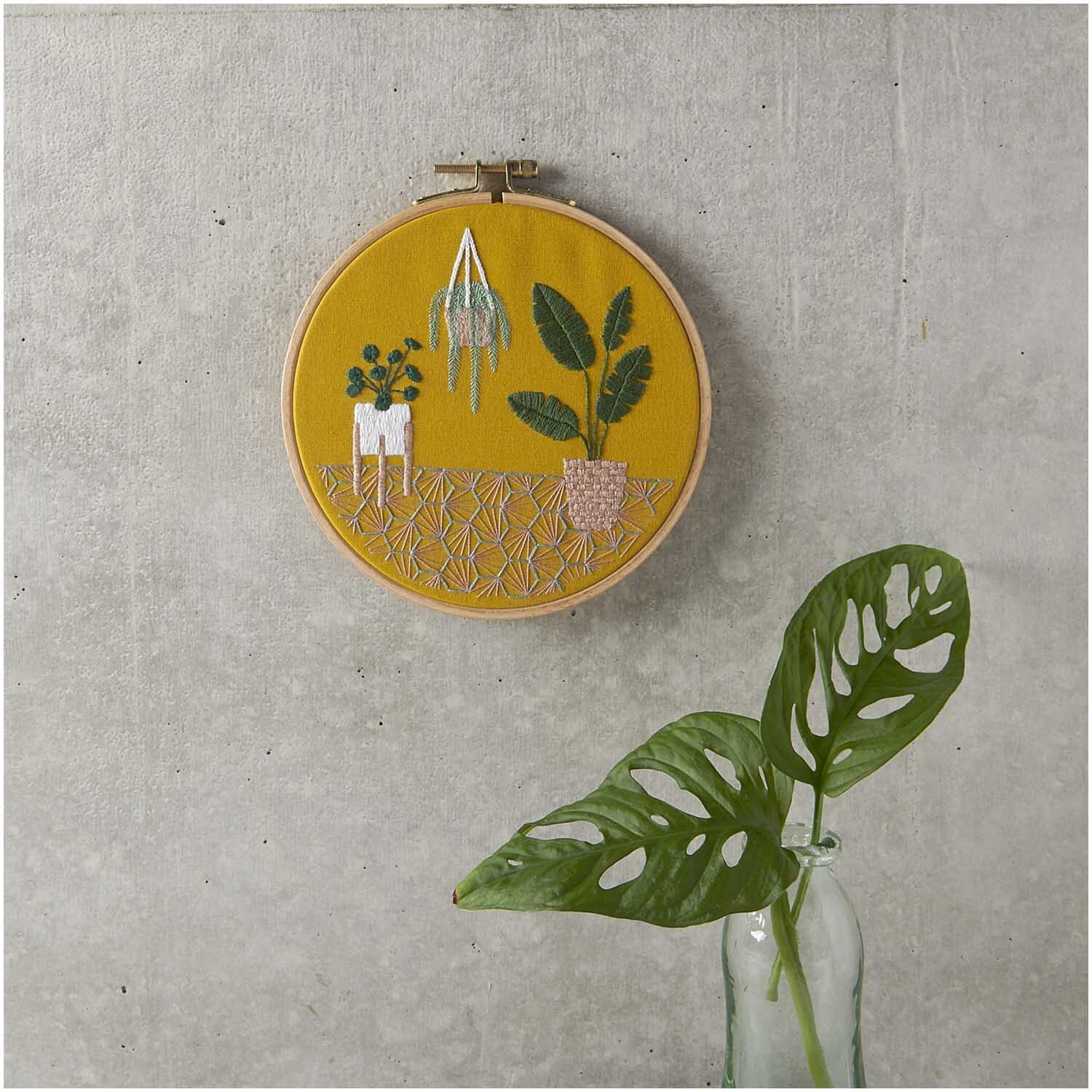Paquet à broder Slow Evenings Urban Jungle image Ø 15,5cm from Rico Design Stickpackung Slow Evenings Urban Jungle Bild Ø 15,5cm zeigt Pflanzenstickerei auf gelbem Stoff, aufgehängt an grauer Wand.