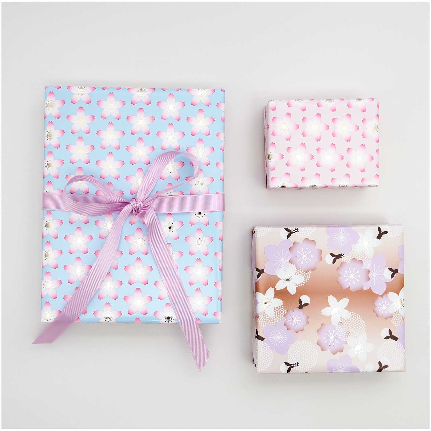 Geschenkpapier Kirschblüten Pink 70cm 2m 80g/m² von Rico Design Geschenkpapier Kirschblüten Pink: Drei Geschenke in pastellfarbenem Papier mit floralen Mustern, verziert mit einer rosa Schleife.