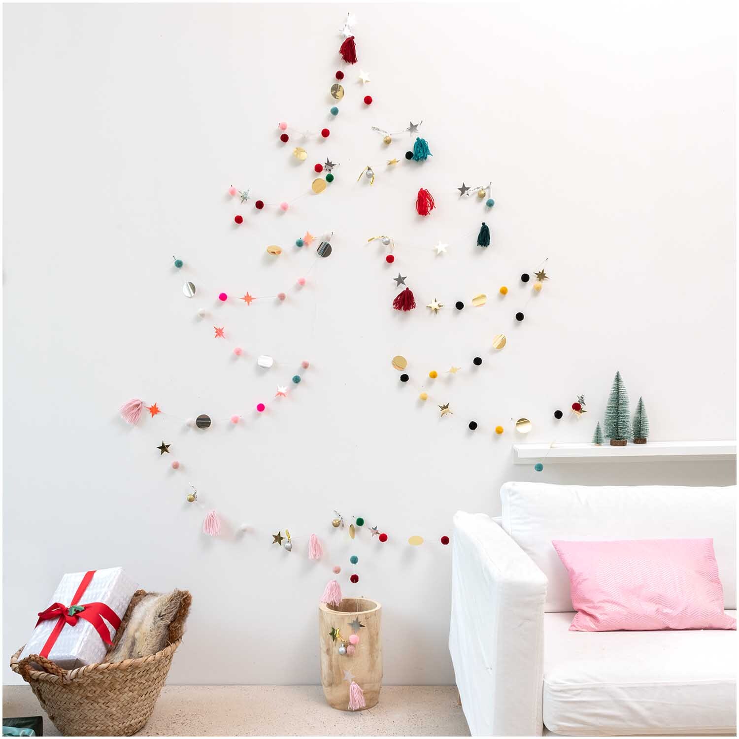 Quasten-Girlande rosa-pastell 2,2m von Rico Design Quasten-Girlande rosa-pastell 2,2m dekoriert eine Wand in Form eines Weihnachtsbaums, umgeben von Geschenken und einem Sofa.