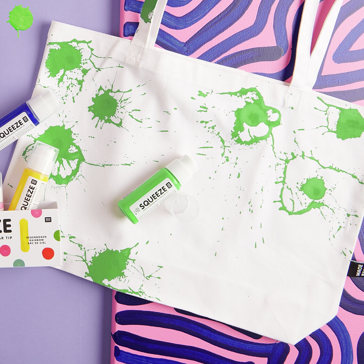 Squeeze Acrylic Marker Set Basic Rainbow 5x60ml von Rico Design Squeeze Acrylic Marker 60ml: Stofftasche mit grünen Farbspritzern, umgeben von bunten Acrylmarkern. Ideal für kreative Designs.