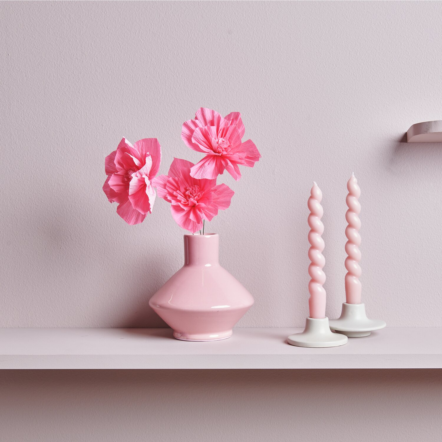 Krepppapier Blüten in Neonpink von Rico Design in Vase, daneben zwei gedrehte Kerzen.