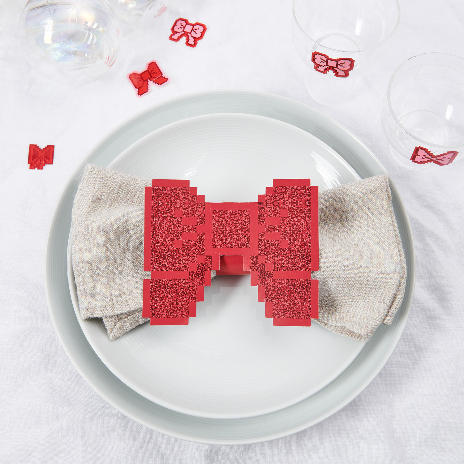 Paper Poetry Serviettenringe Schleifen Pixel 12x10cm 6 Stück von Rico Design Paper Poetry Serviettenringe Schleifen Pixel in Rot auf einem gedeckten Tisch mit weißem Geschirr und Leinenserviette, 6er-Set von Rico Design.