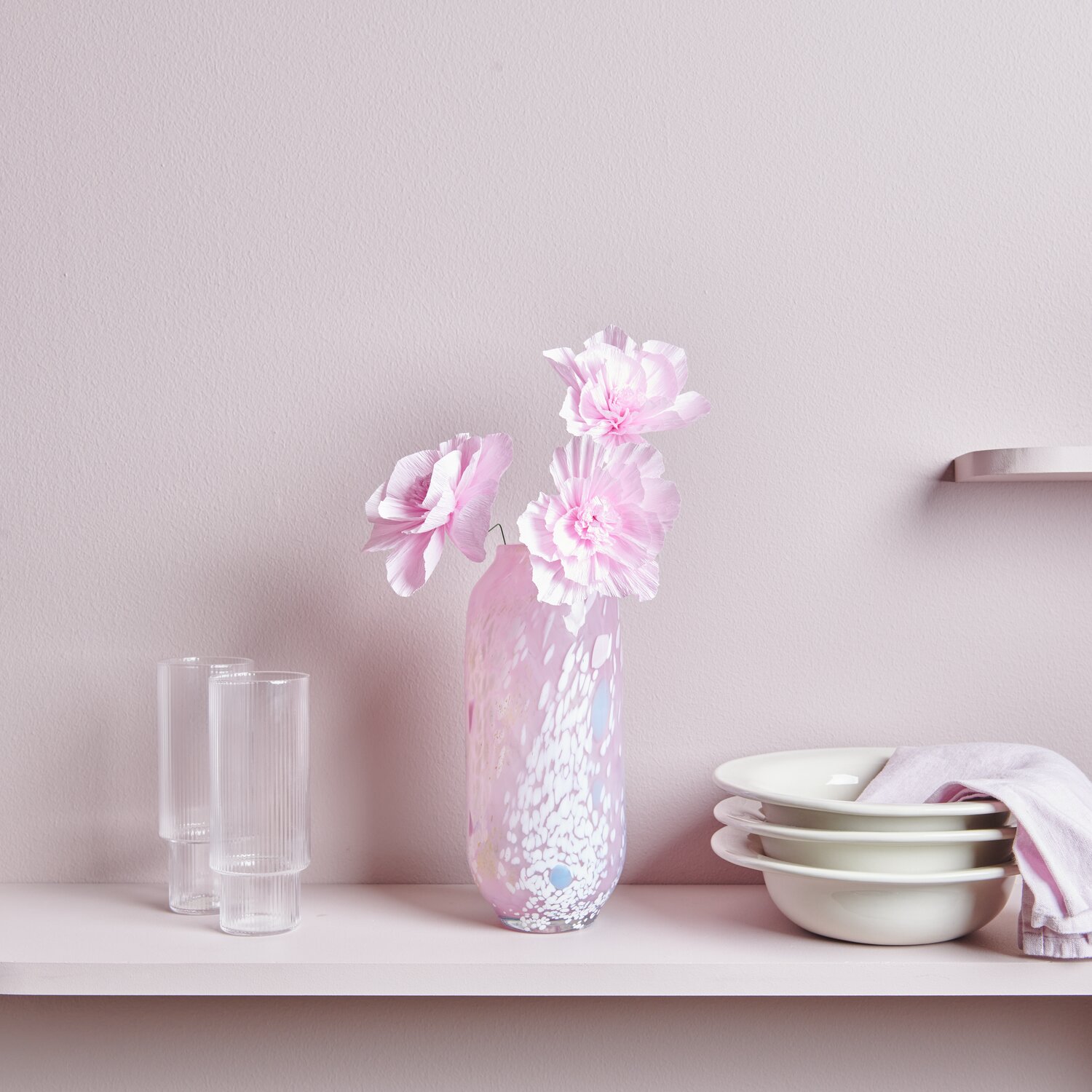 Krepppapier Blüten in einer Vase auf einem Regal, umgeben von Gläsern und Schalen, in sanftem Rosa dekoriert.