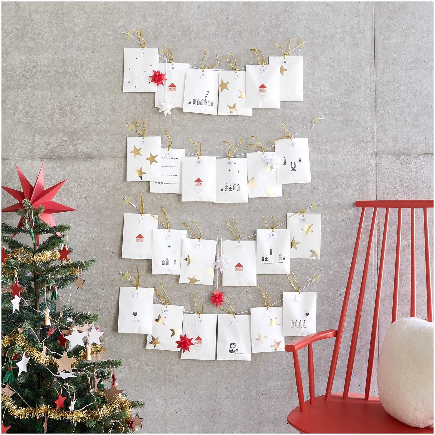 Paper Poetry Adventskalenderanhänger, 24 Stück, dekoriert mit goldenen Sternen an einer Wand. Links steht ein geschmückter Weihnachtsbaum.