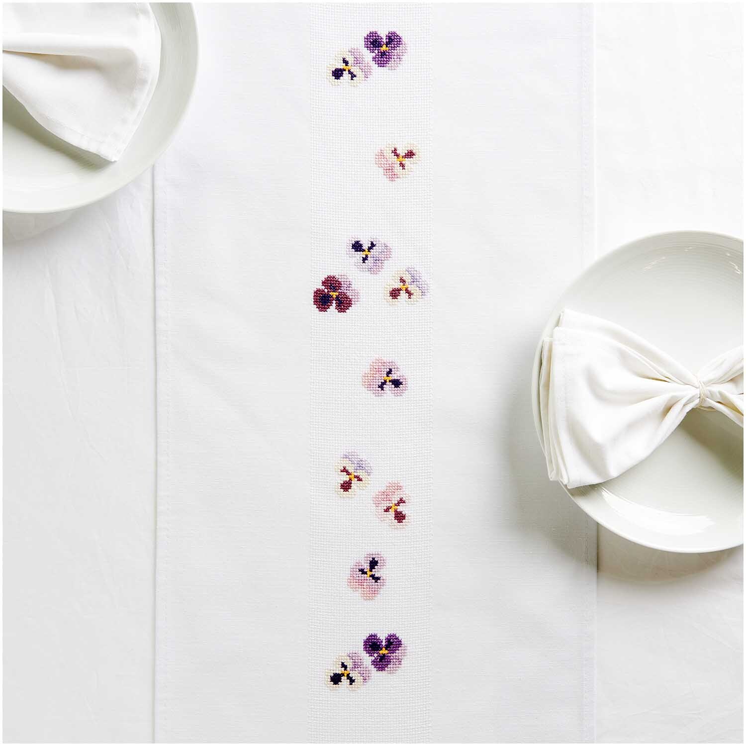 Paquet à broder ruban de table Pensées 30x160cm from Rico Design Stickpackung Tischband Stiefmütterchen von Rico Design, dekoriert mit zarten Blumenstickereien auf weißem Stoff, ideal für elegante Tischdekorationen.