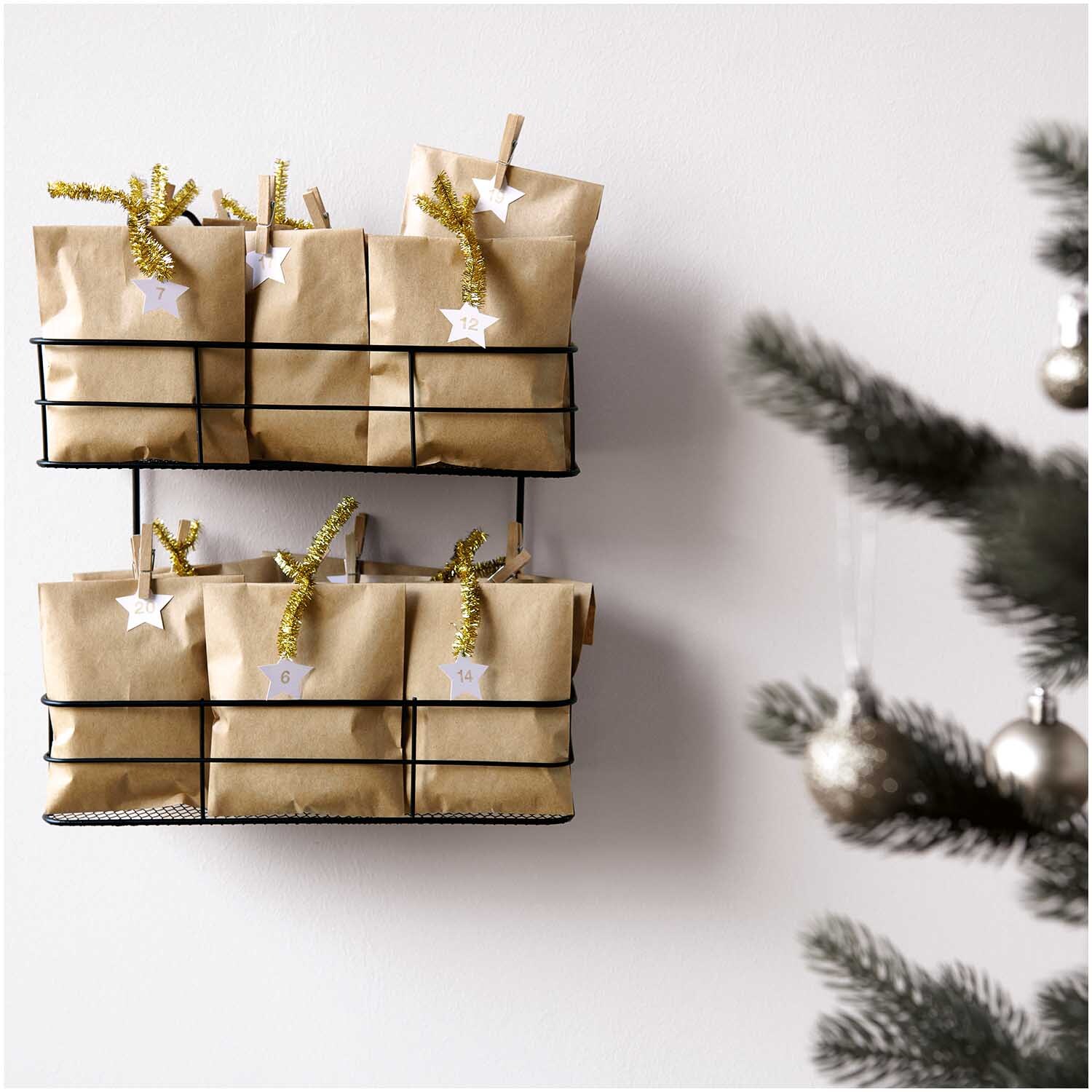 Paper Poetry Adventskalenderanhänger Sterne, dekoriert an braunen Geschenktüten in einem Regal, mit weihnachtlichem Baum im Hintergrund.