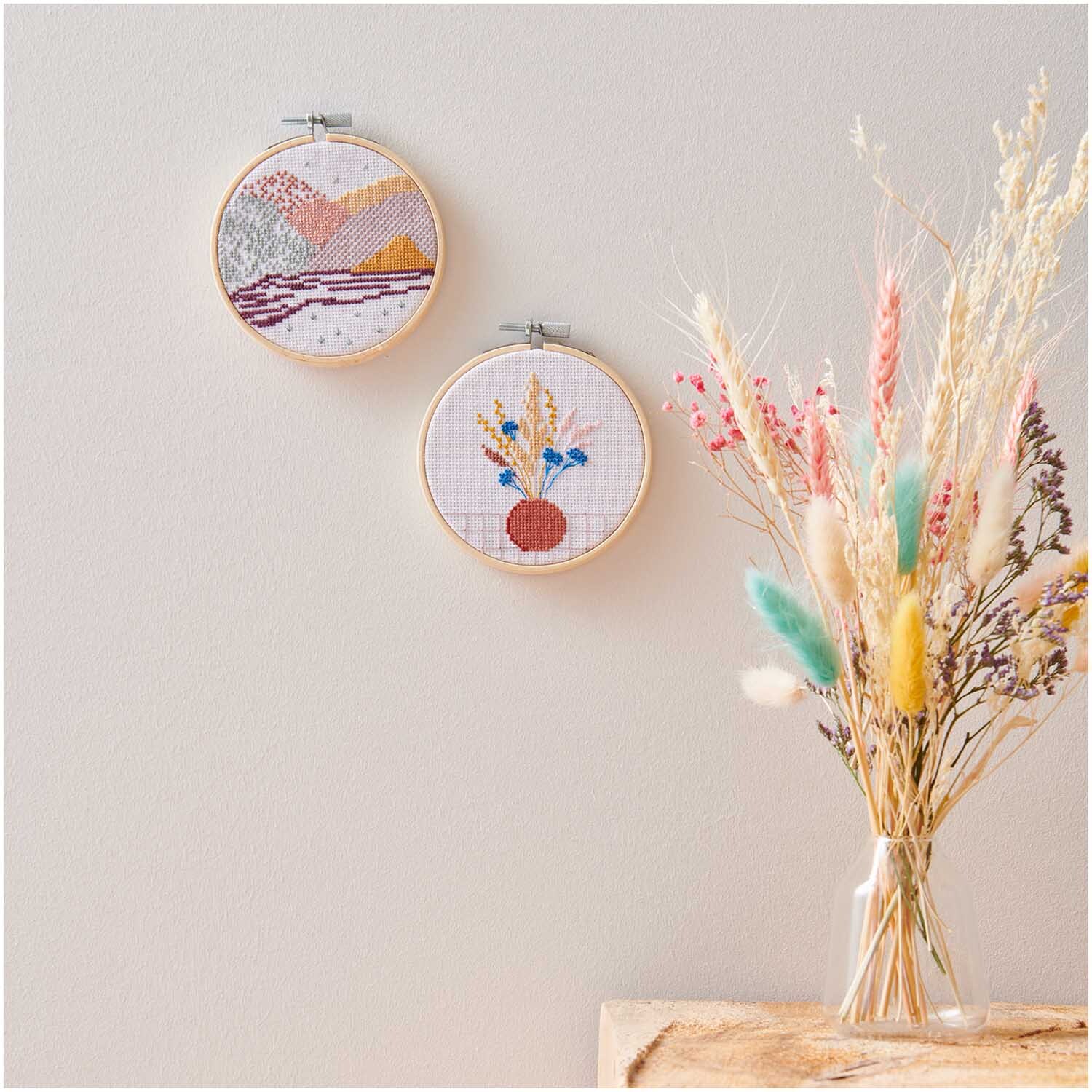 Kit de broderie Mini Kit Fleurs séchées Image Ø 10cm from Rico Design Stickpackung Mini Kit Trockenblumen Bild Ø 10cm zeigt bunte Stickereien an einer Wand. Daneben steht ein Strauß Trockenblumen in einer Vase.