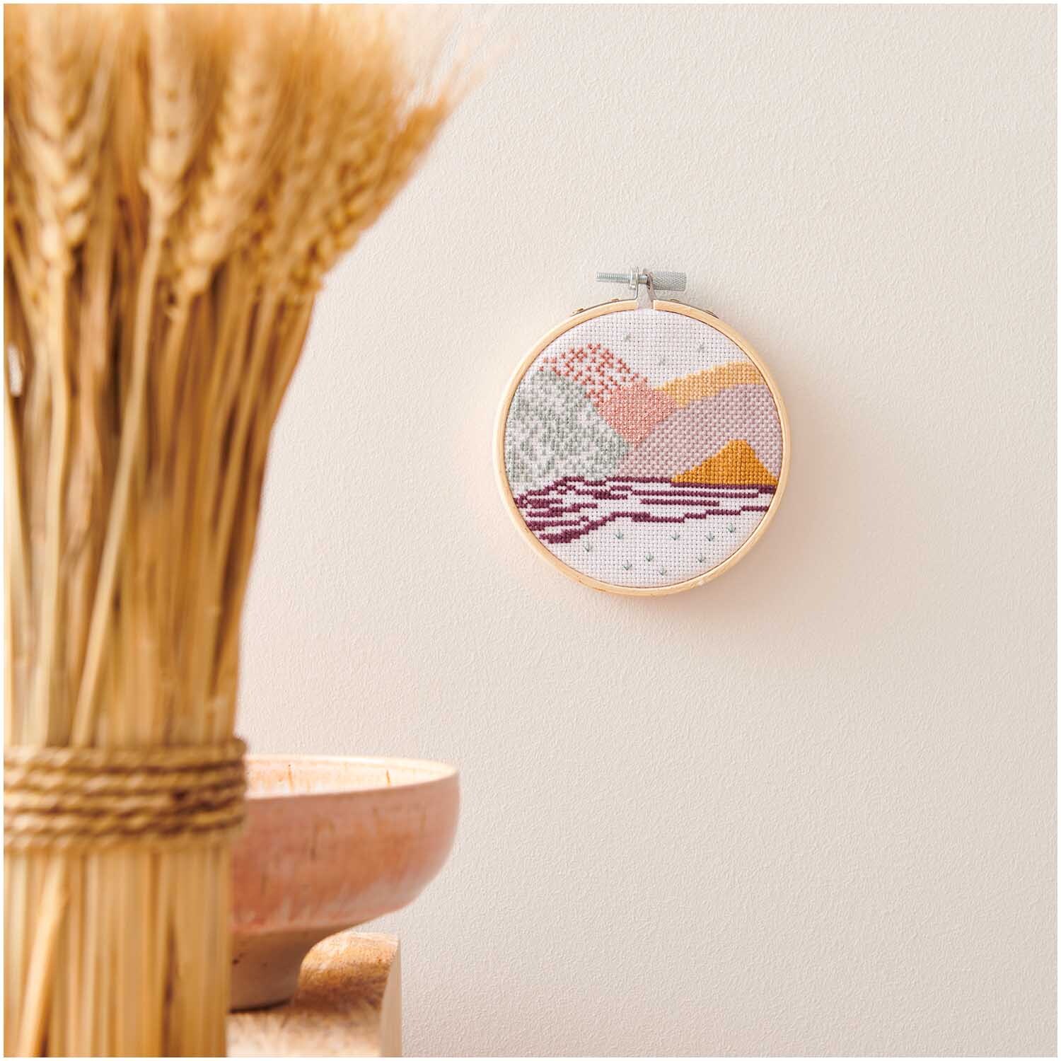 Mini Kit de broderie Paysage Image Ø 10cm from Rico Design Stickpackung Mini Kit Landschaft Bild Ø 10cm an einer Wand, mit warmen Farbtönen und Holzrahmen.