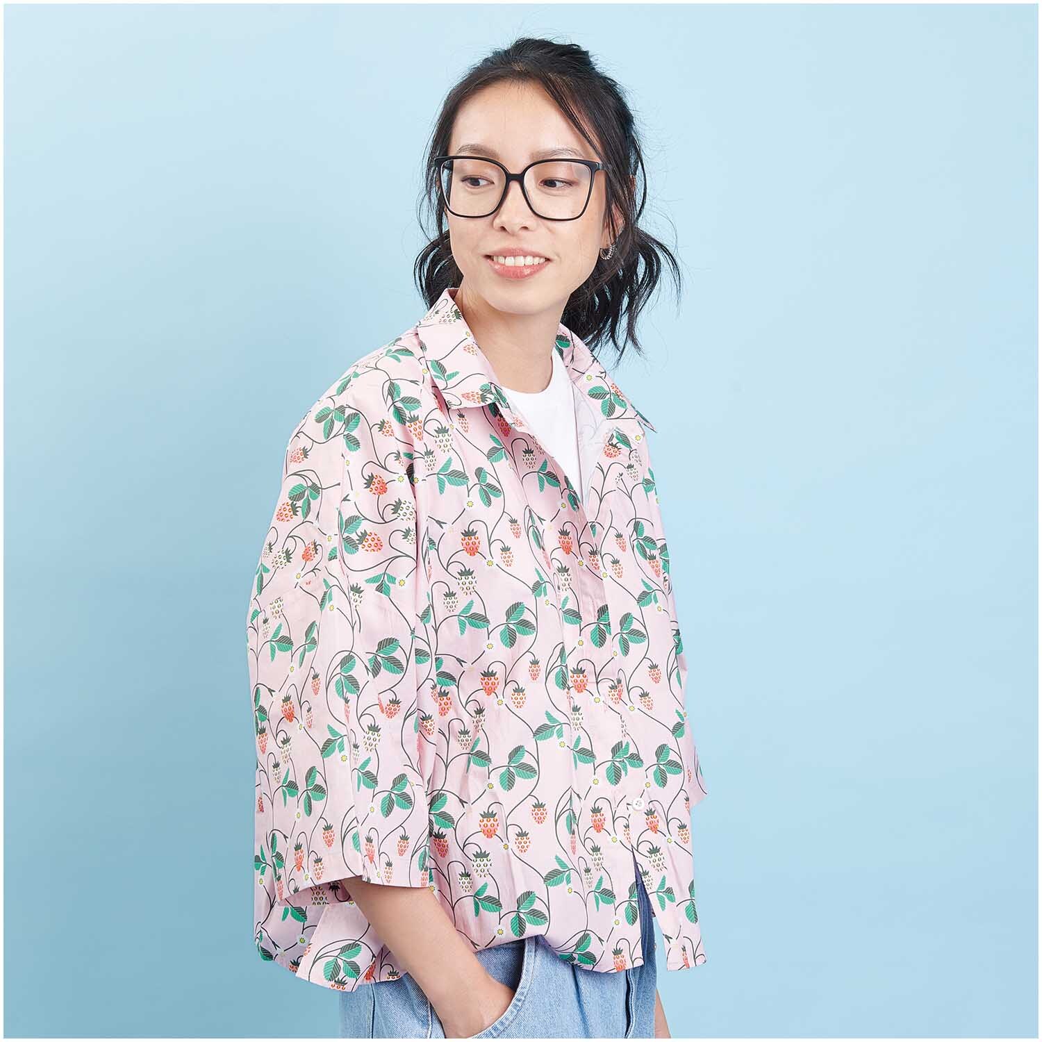 Meterware Baumwoll-Popelin rosa Erdbeeren von Rico Design: Frau trägt ein Hemd mit Erdbeerprint vor blauem Hintergrund.