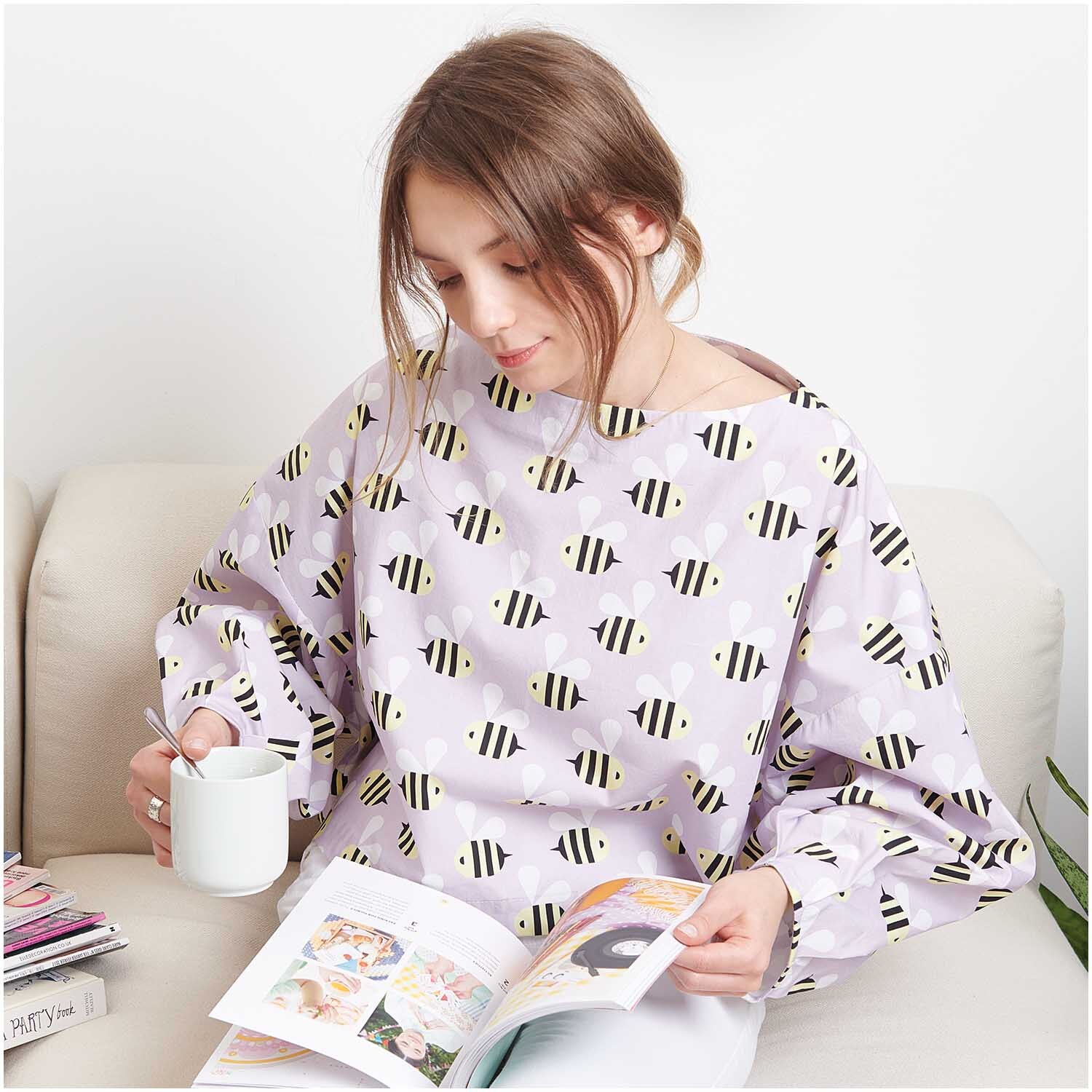 Stoffabschnitt Baumwoll-Popelin flieder Bienen 50x140cm von Rico Design Stoffabschnitt Baumwoll-Popelin flieder Bienen: Frau in fliederfarbenem Oberteil mit Bienenmuster, liest ein Buch und hält eine Tasse.