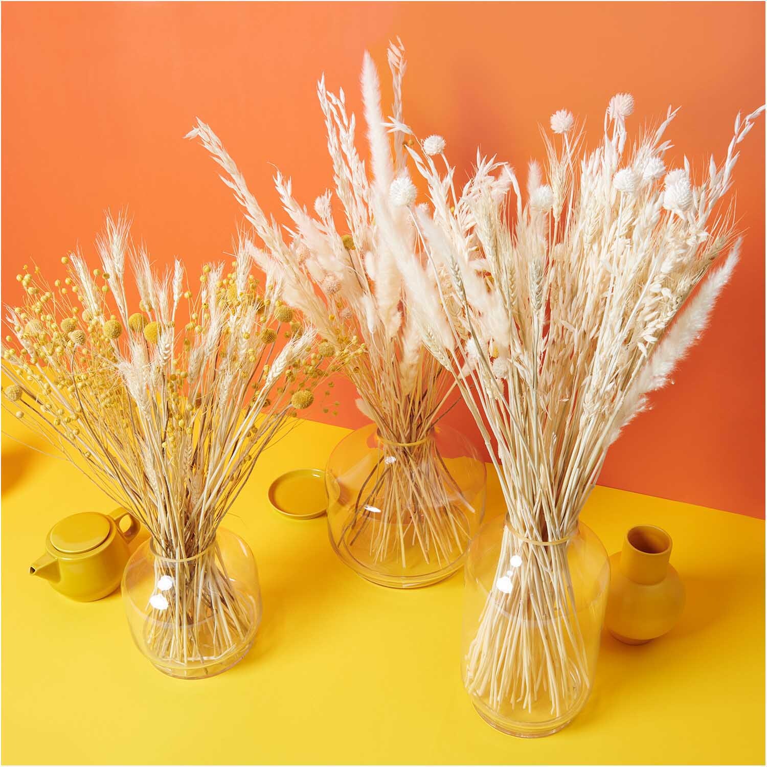 Getrockneter Weizen natur 60-70cm 50 Stück von Rico Design Getrockneter Weizen natur 60-70cm 50 Stück in Glasvasen auf gelbem Tisch vor orangefarbener Wand.