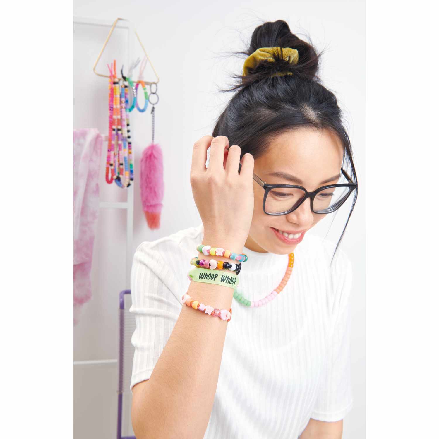 itoshii - Ponii Beads Bügelmotiv Schriftzüge 8 Designs von Rico Design Itoshii - Ponii Beads Bügelmotiv Schriftzüge 8 Designs: Frau mit bunten Armbändern und Brille, lächelnd in einem kreativen Raum.