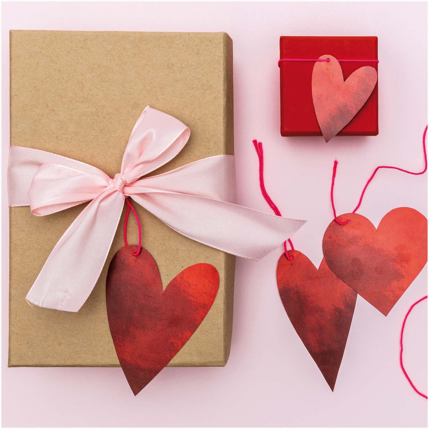 Paper Poetry Papieranhänger in Herzform, dekorativ an Geschenkboxen mit rosa Schleife und rotem Etui arrangiert.