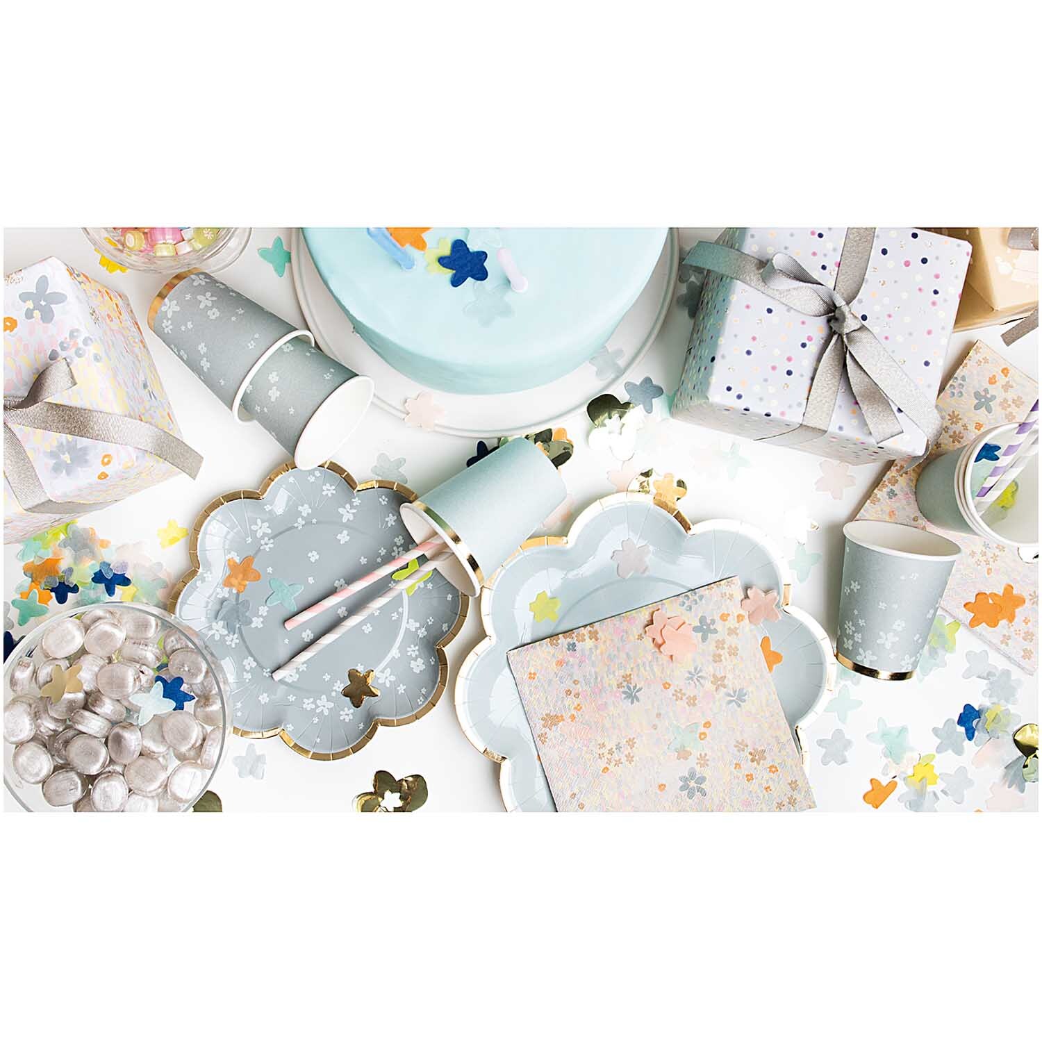 Konfetti Blüten blau Mix 20g von Rico Design Konfetti Blüten blau Mix 20g: Festliche Tischdekoration mit blauen Blütenkonfetti von Rico Design, umgeben von Partygeschirr und Geschenken.