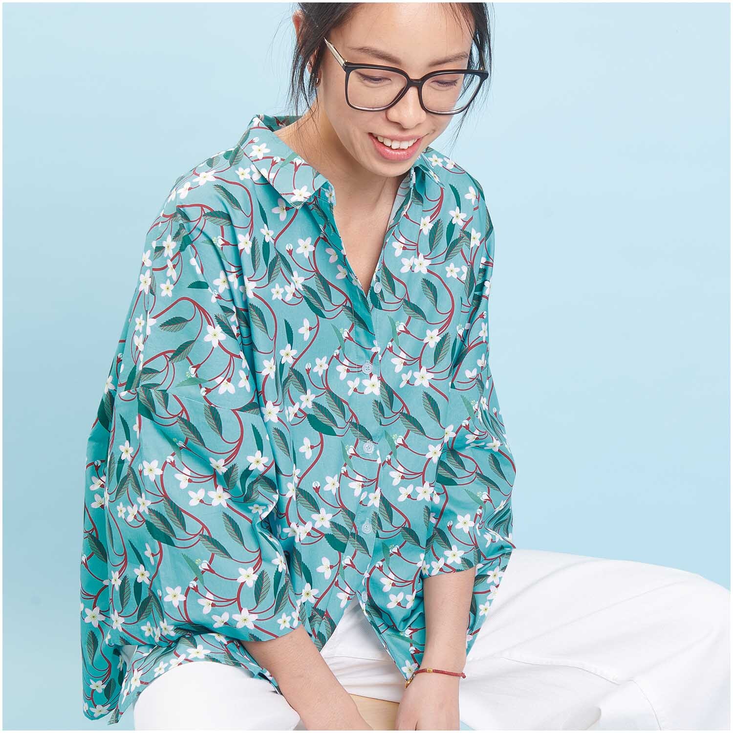 Meterware Baumwoll-Popelin türkis Kirschblüten von Rico Design Meterware Baumwoll-Popelin türkis Kirschblüten von Rico Design: Frau trägt ein türkisfarbenes Hemd mit Kirschblütenmuster, sitzt lächelnd vor blauem Hintergrund.