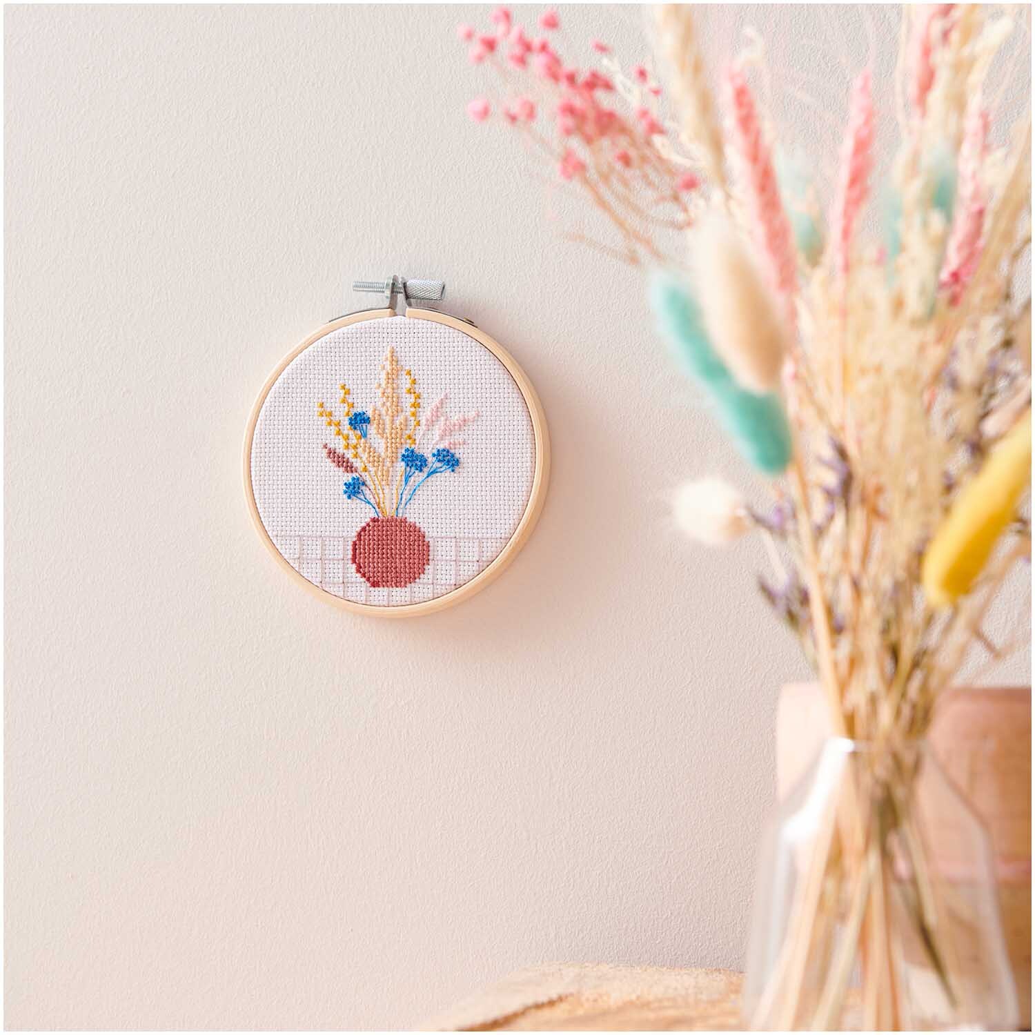 Kit de broderie Mini Kit Fleurs séchées Image Ø 10cm from Rico Design Stickpackung Mini Kit Trockenblumen Bild Ø 10cm zeigt bunte Blumen in einem Stickrahmen an der Wand.