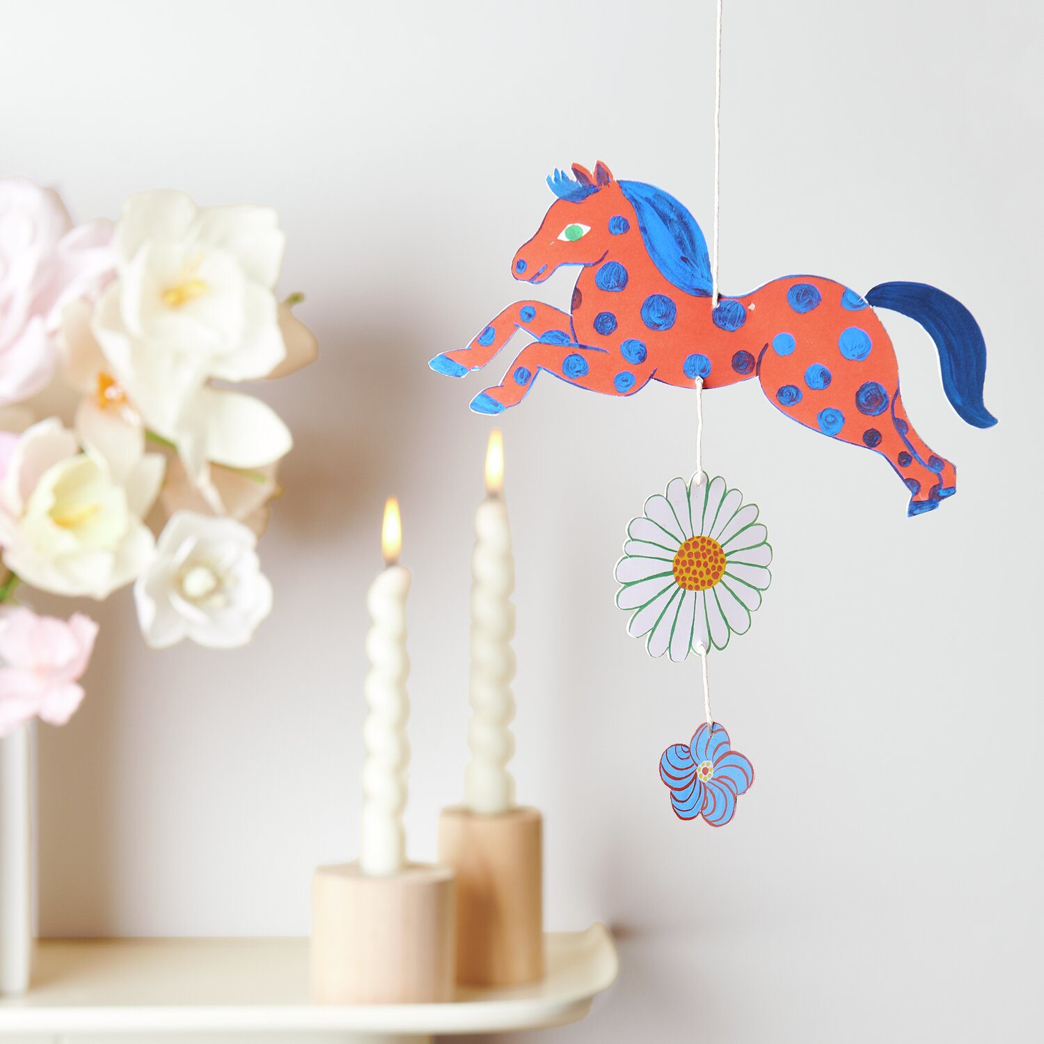 Bastelpapierblock Midsommar Magic 23 Blatt von Rico Design Bastelpapierblock Midsommar Magic: Buntes Pferde-Mobile mit Blumen, dekorativ neben weißen Kerzen und Blumenarrangement.