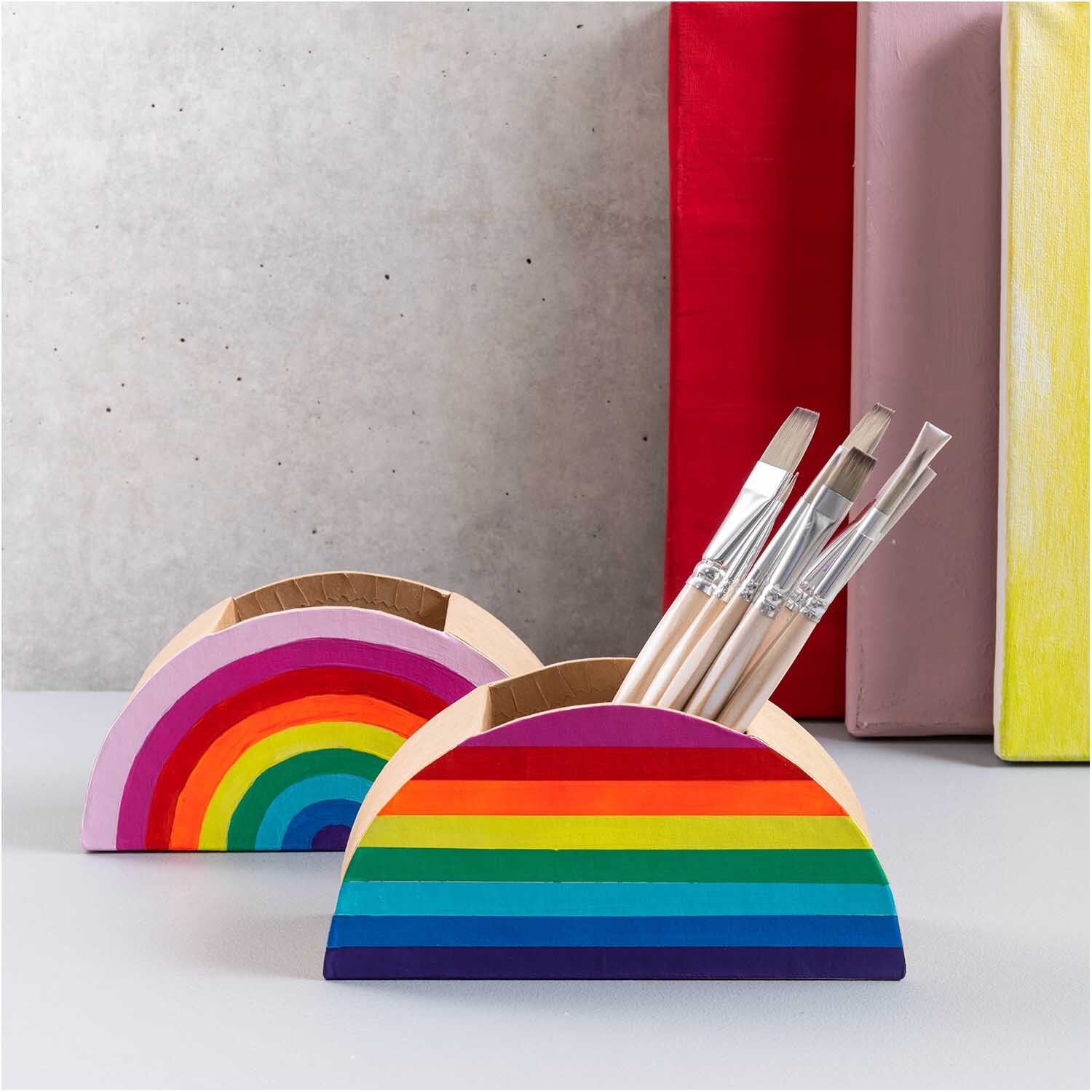 Stiftehalter aus Pappe halbrund 18x9x5cm von Rico Design in Regenbogenfarben, gefüllt mit Pinseln, vor buntem Hintergrund.