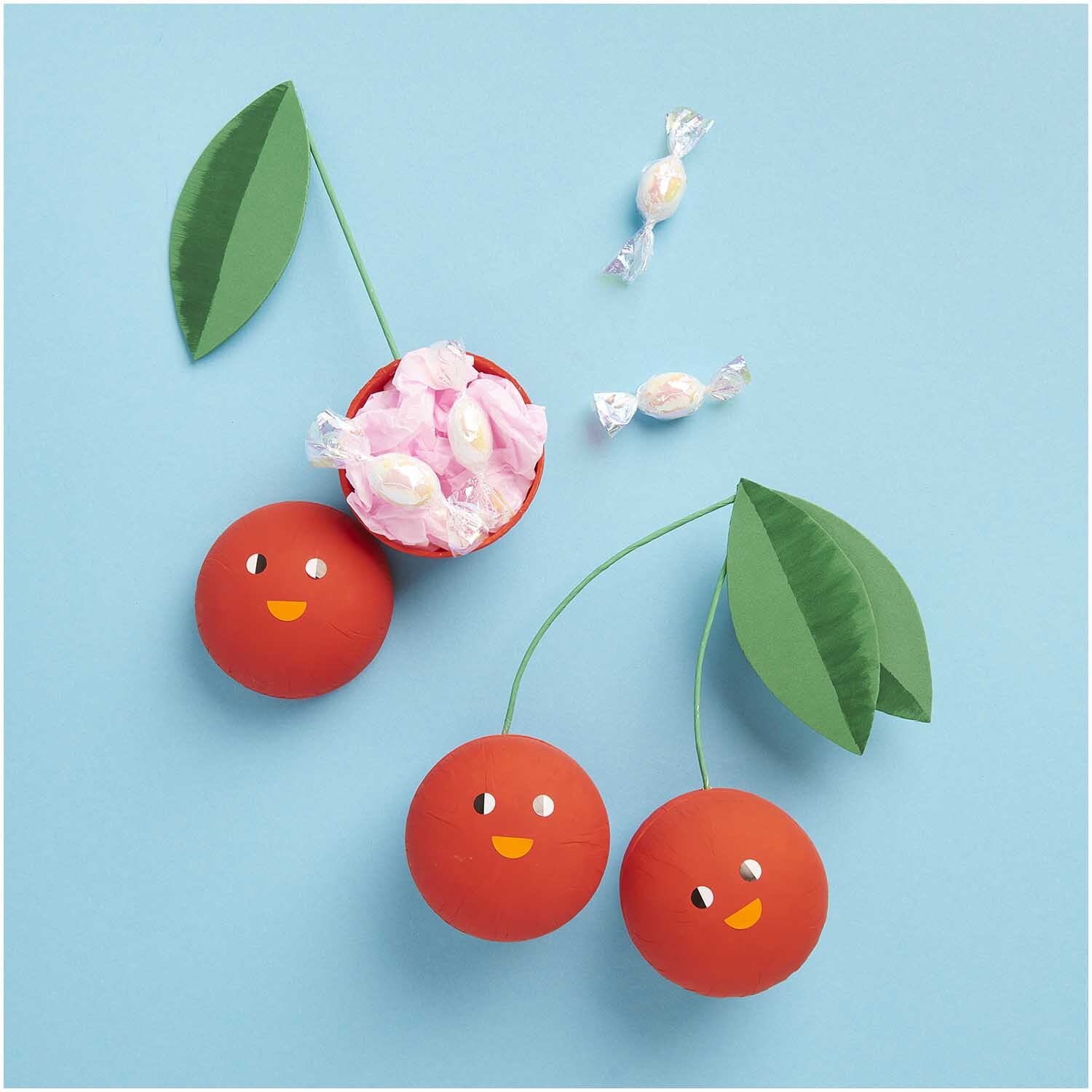 Pappmaché Kugel natur 8cm von Rico Design: Drei rote Kugeln mit Gesichtern und grünen Blättern auf blauem Hintergrund.