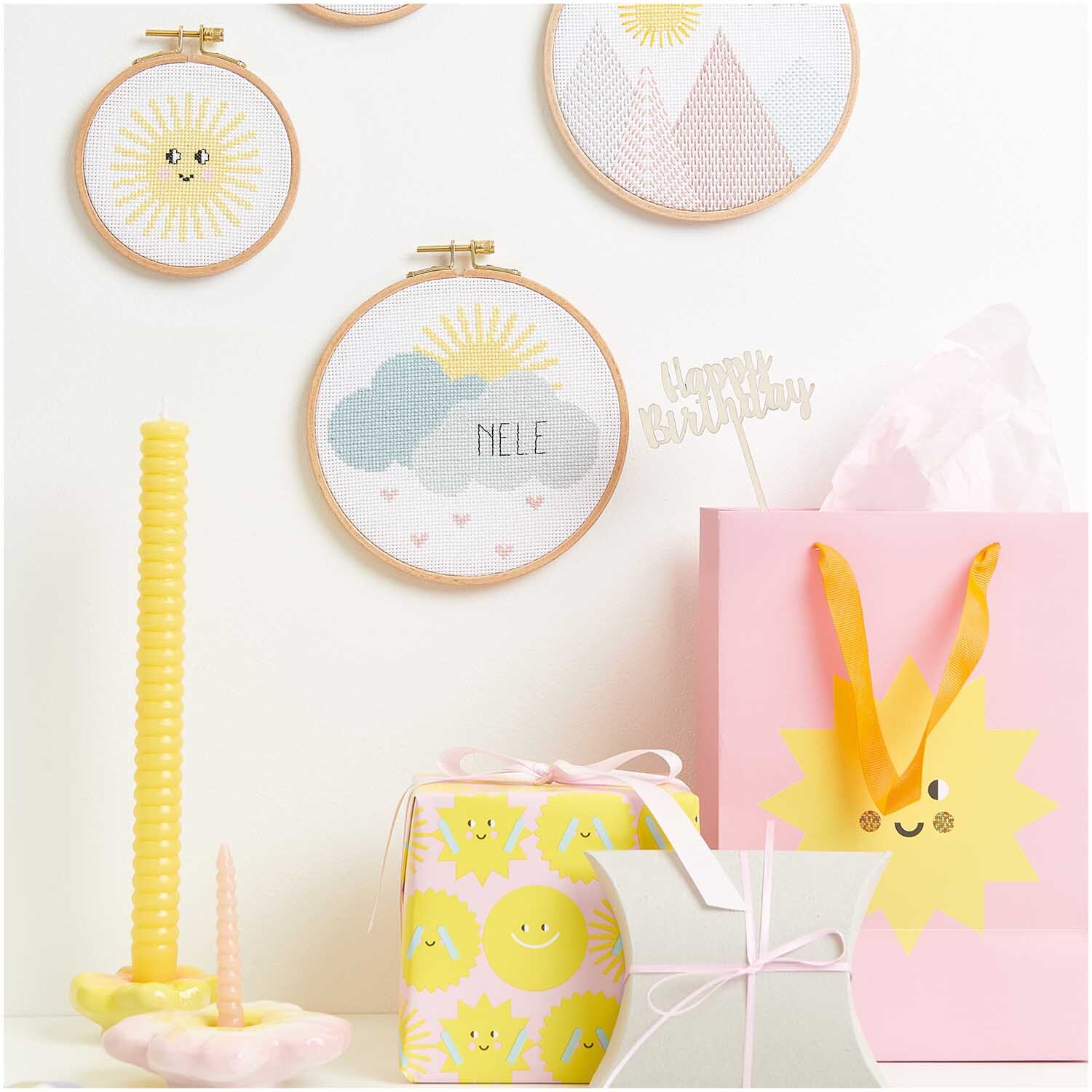 Kit de broderie image de naissance nuages from Rico Design Stickpackung Geburtsbild Wolken von Rico Design zeigt bunte Stickrahmen mit Wolkenmotiv, umgeben von fröhlichen Geschenken und Dekorationen.