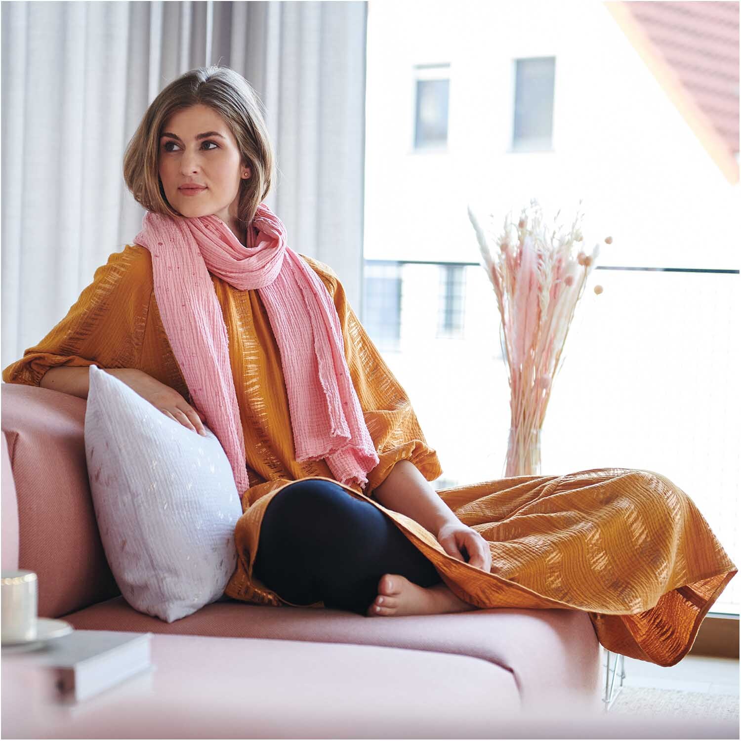 Meterware Krinkelmusselin hellgrau Hot Foil von Rico Design Meterware Krinkelmusselin hellgrau Hot Foil: Frau in gelbem Kleid mit rosa Schal auf rosa Sofa, modernes Wohnzimmer.