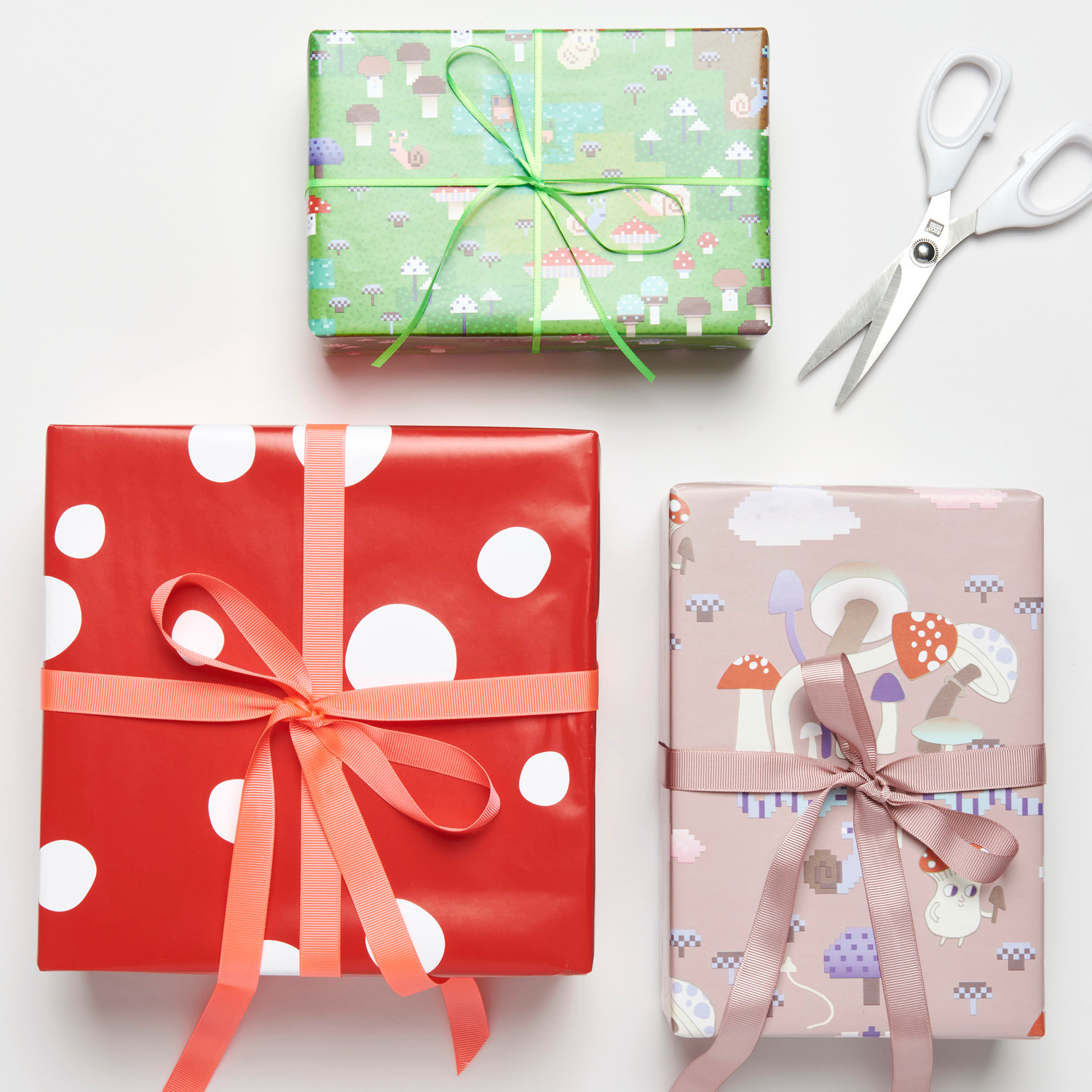 Geschenkpapier Shrooom Landschaft 70cm 2m 80g/m² von Rico Design Geschenkpapier Shrooom Games, Fliegenpilz-Muster in Rot, Grün und Rosa, dekorativ verpackt mit Schleifen, daneben eine Schere.