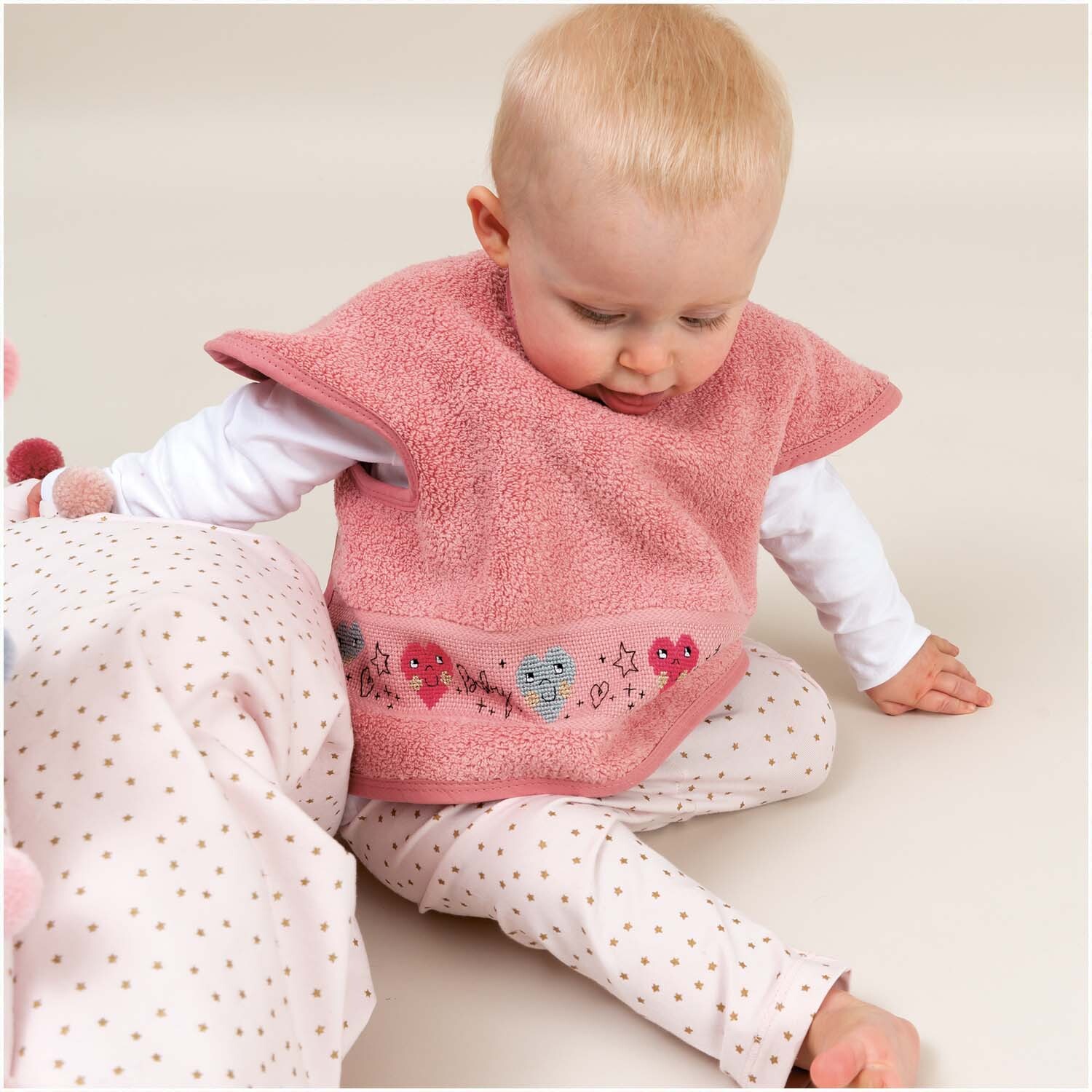 Ärmellätzchen 40x49cm in Hellpink von Rico Design, getragen von einem Baby, das auf dem Boden sitzt.