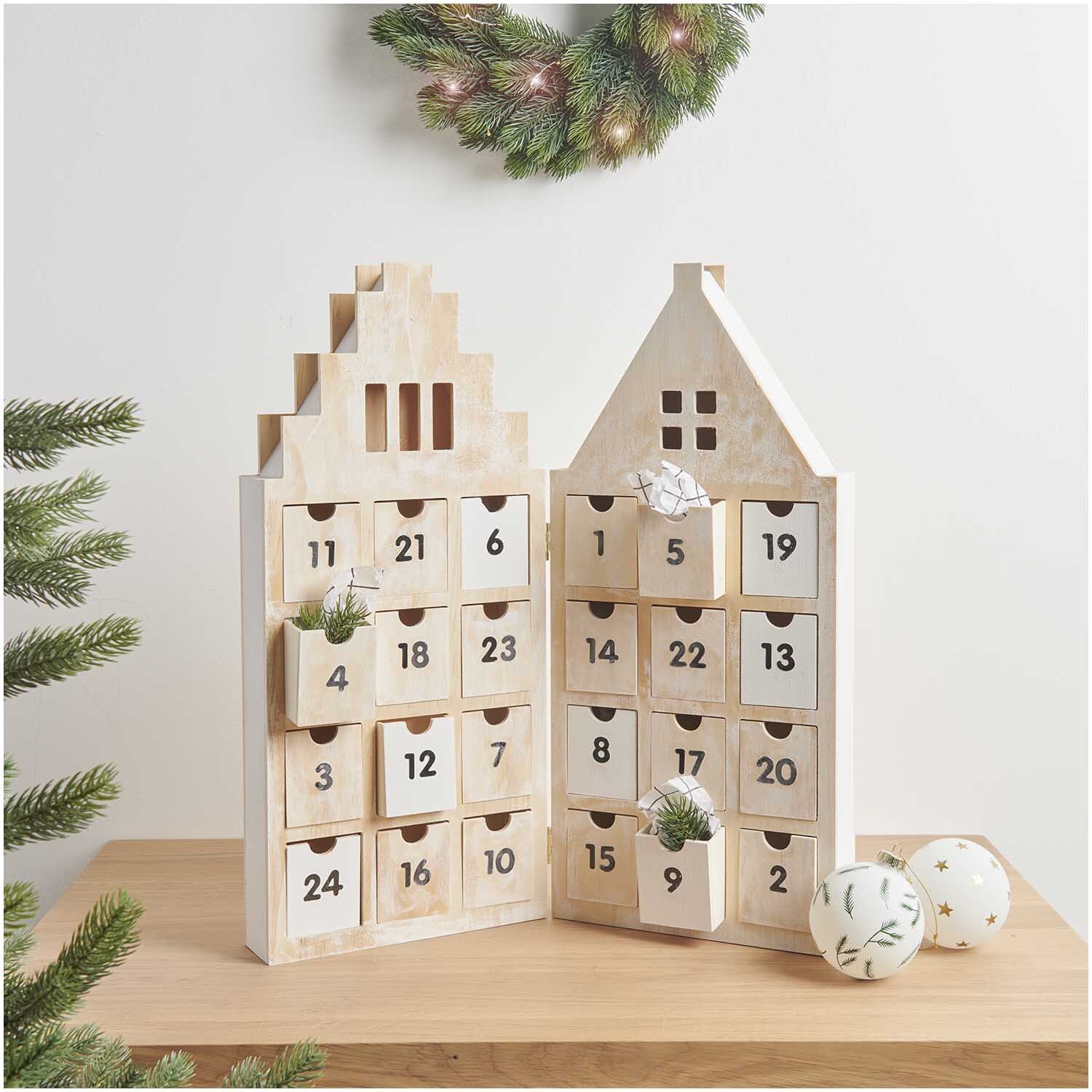 Adventskalender Haus aus Holz 46,5x51x5cm von Rico Design Adventskalender Haus aus Holz mit 24 nummerierten Schubladen, dekoriert mit Tannenzweigen und Kugeln, auf einem Tisch platziert.