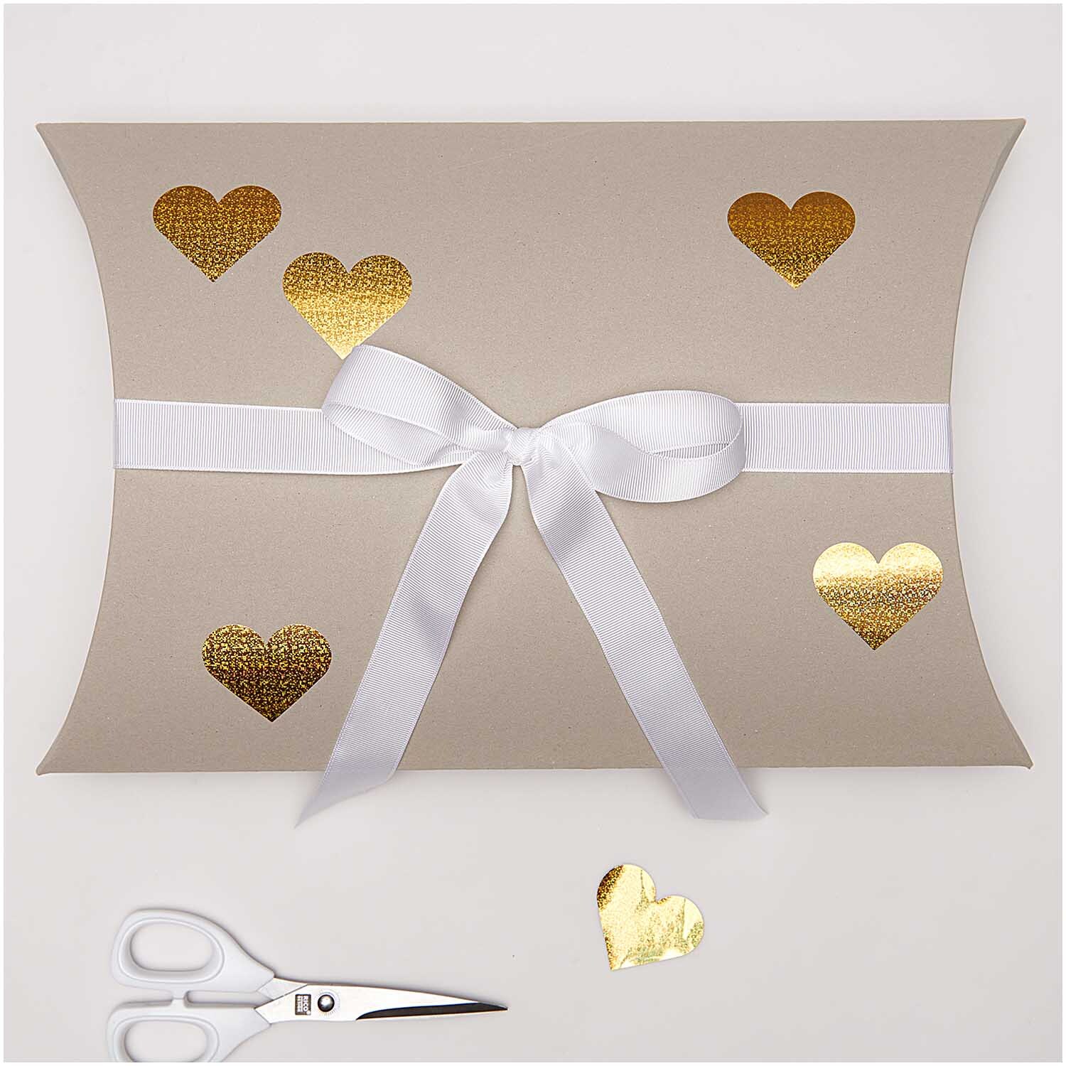 Paper Poetry Sticker Herzen 5cm 120 Stück auf der Rolle Hot Foil Farbe Gold-Holographisch von Rico Design Paper Poetry Sticker Herzen auf Geschenkbox mit goldenen, holographischen Herzen und weißer Schleife. Schere im Vordergrund.